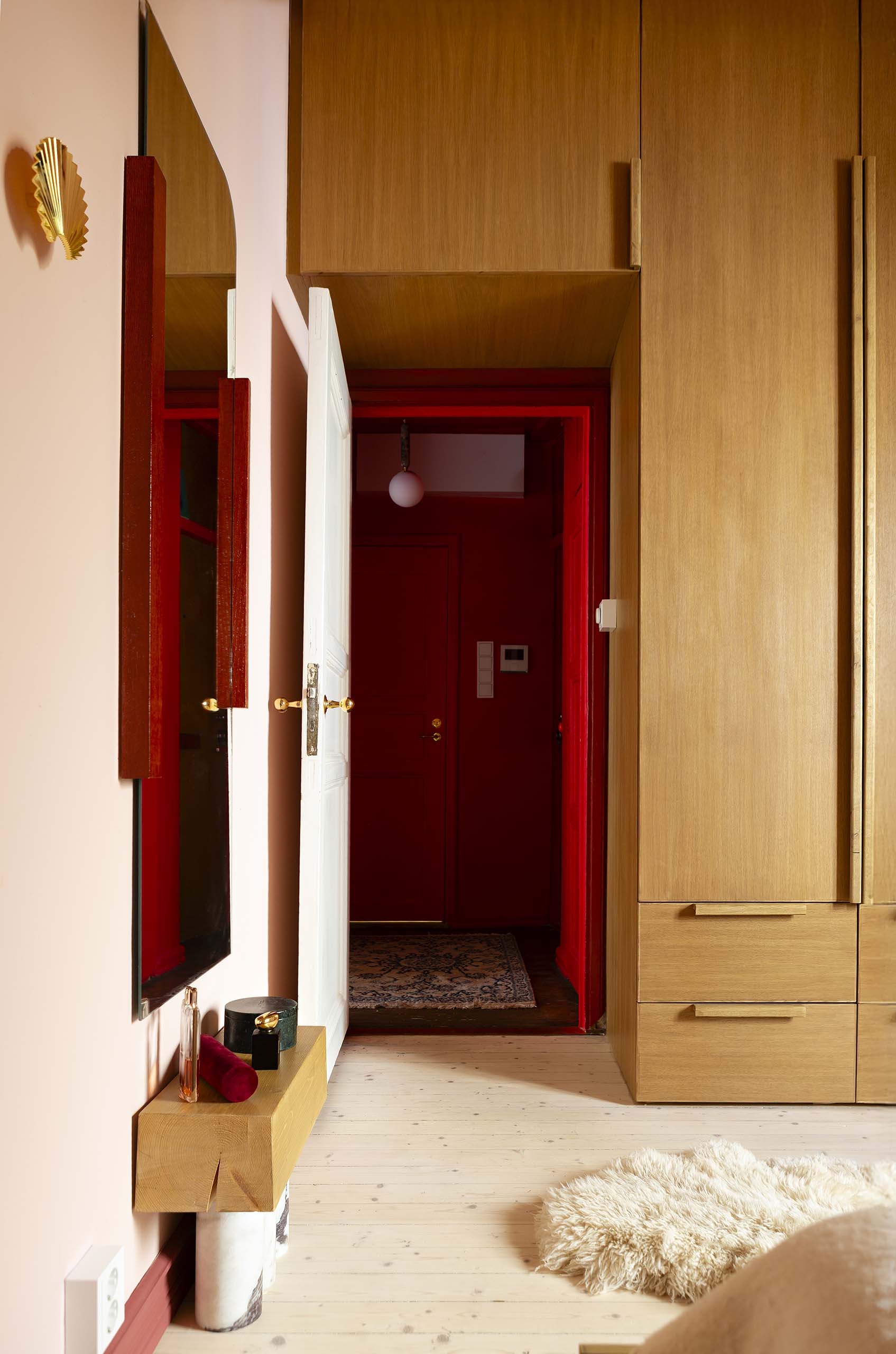 Interior view of a room featuring light wood cabinets, a white door open to a hallway with red walls, a wall mirror, and a cream-colored fluffy rug on pale wooden floor.