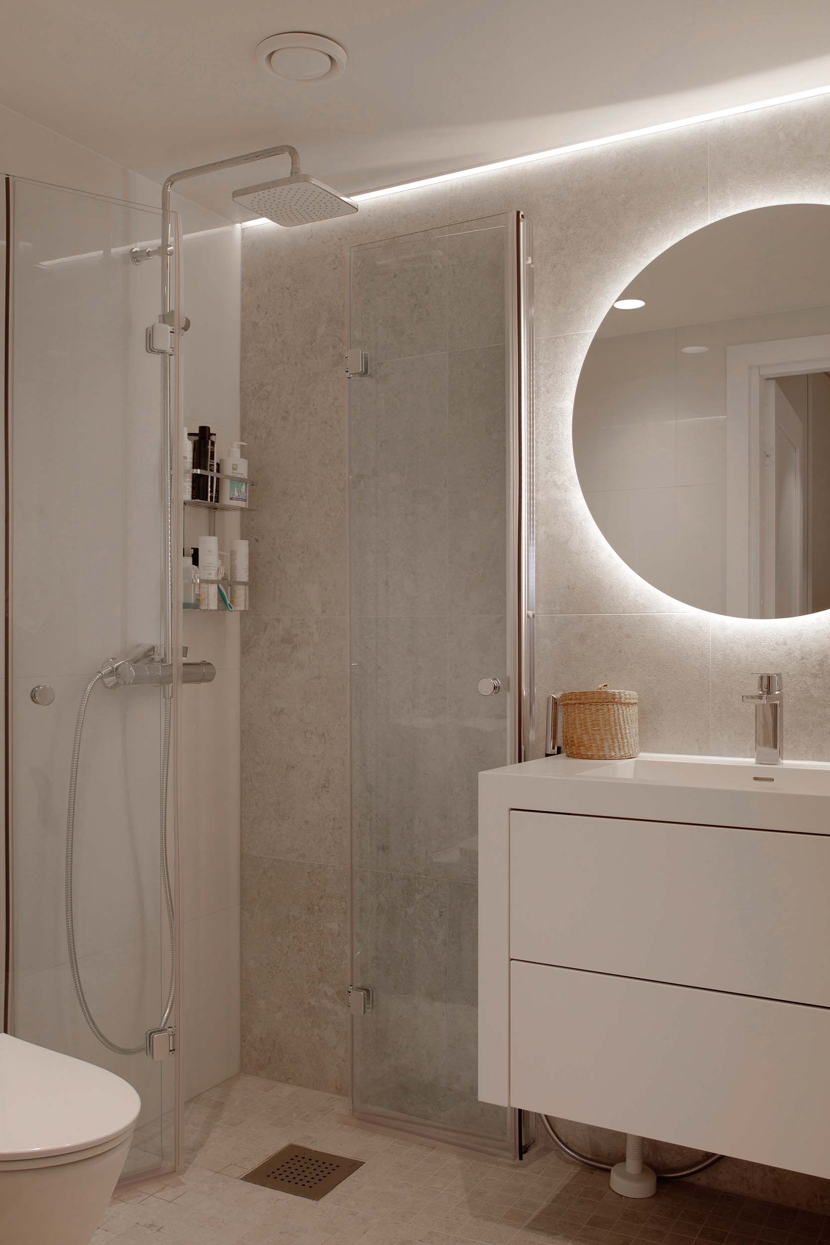 Modern bathroom with glass-enclosed shower, white vanity with round illuminated mirror, and beige tiled walls and floor.