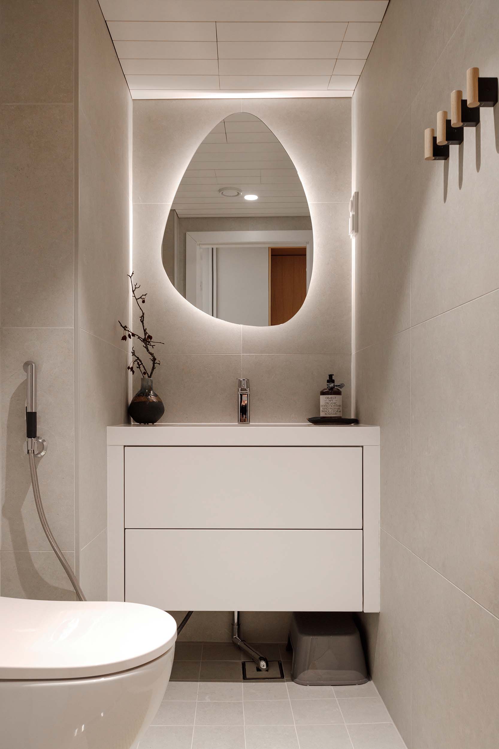 Minimalist bathroom with a white wall-mounted vanity, irregularly shaped backlit mirror, small vase with branches, liquid soap dispenser, and a wall-mounted toilet.