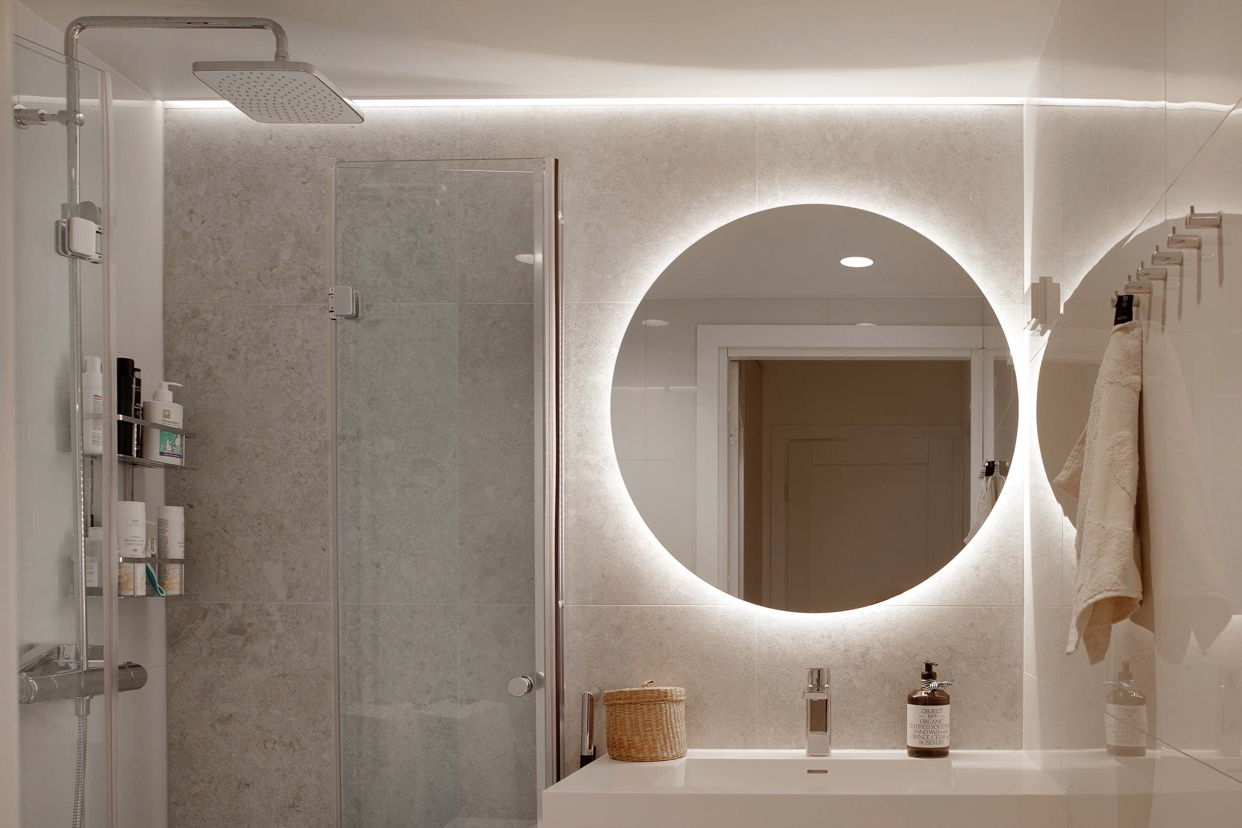 Modern bathroom with a large round backlit mirror above a white sink, glass shower door, and wall-mounted towel hooks holding a beige towel.