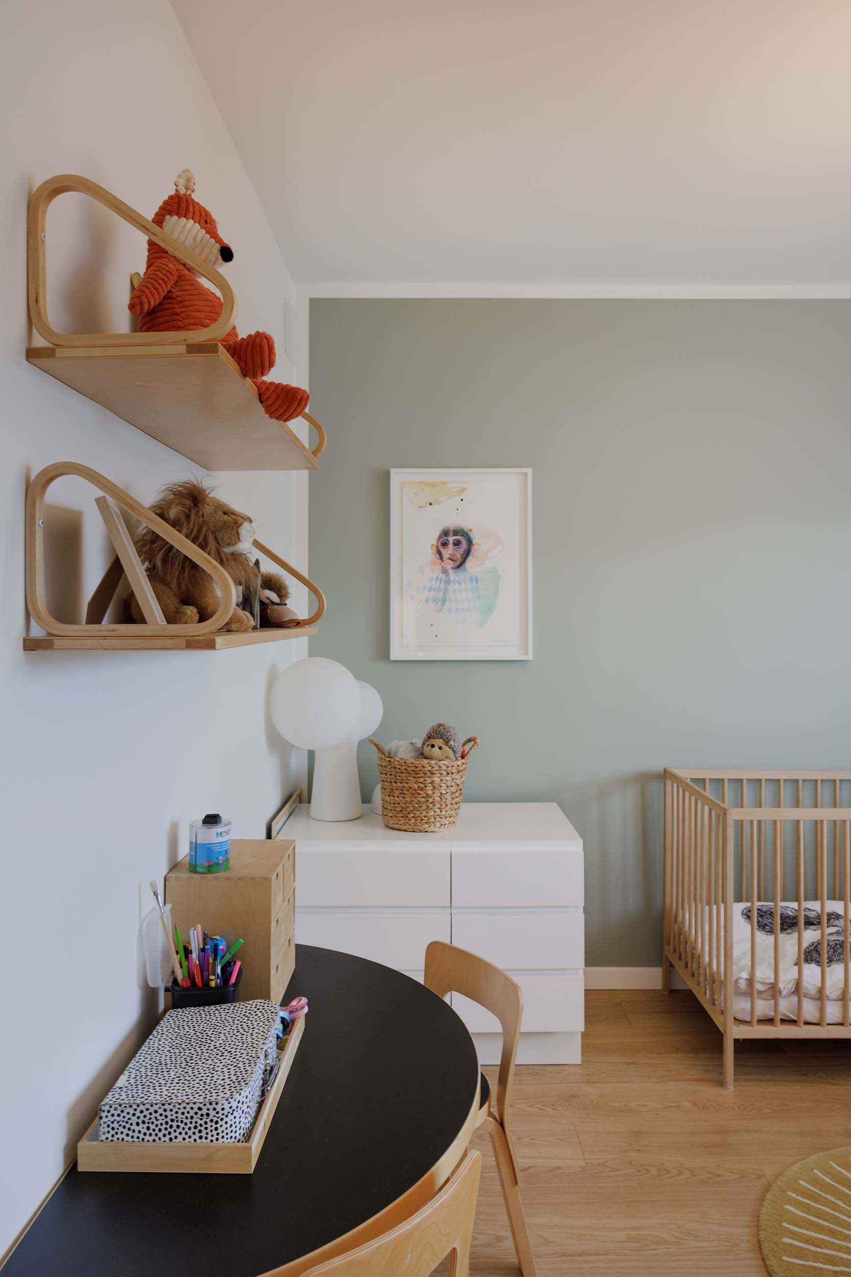 Minimalist nursery with light wood crib, white dresser topped with woven basket and lamp, and two wooden shelves holding stuffed animals on white and green walls.