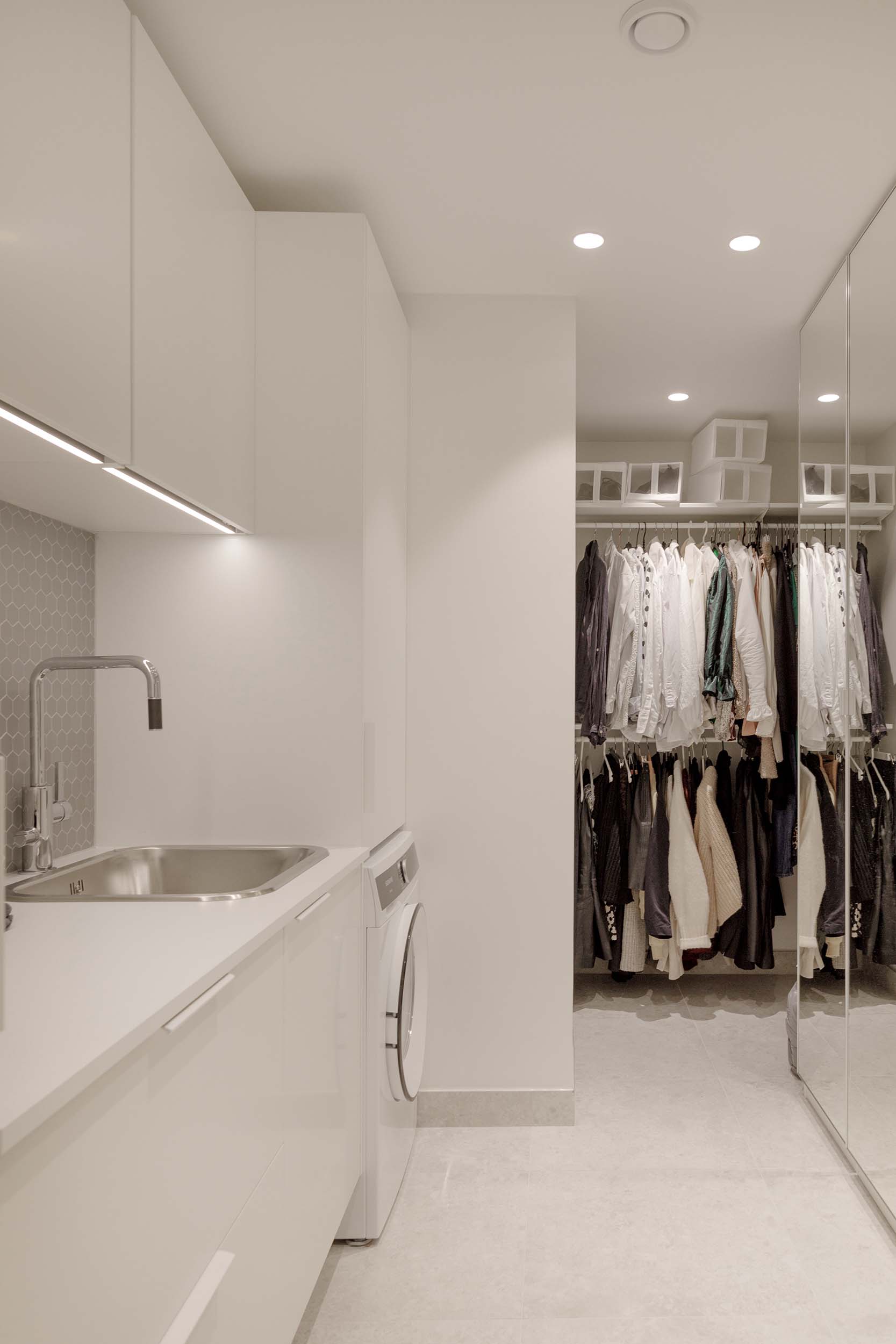 Modern laundry room with white cabinets, stainless steel sink, washing machine, and a closet with clothes hanging on rods.