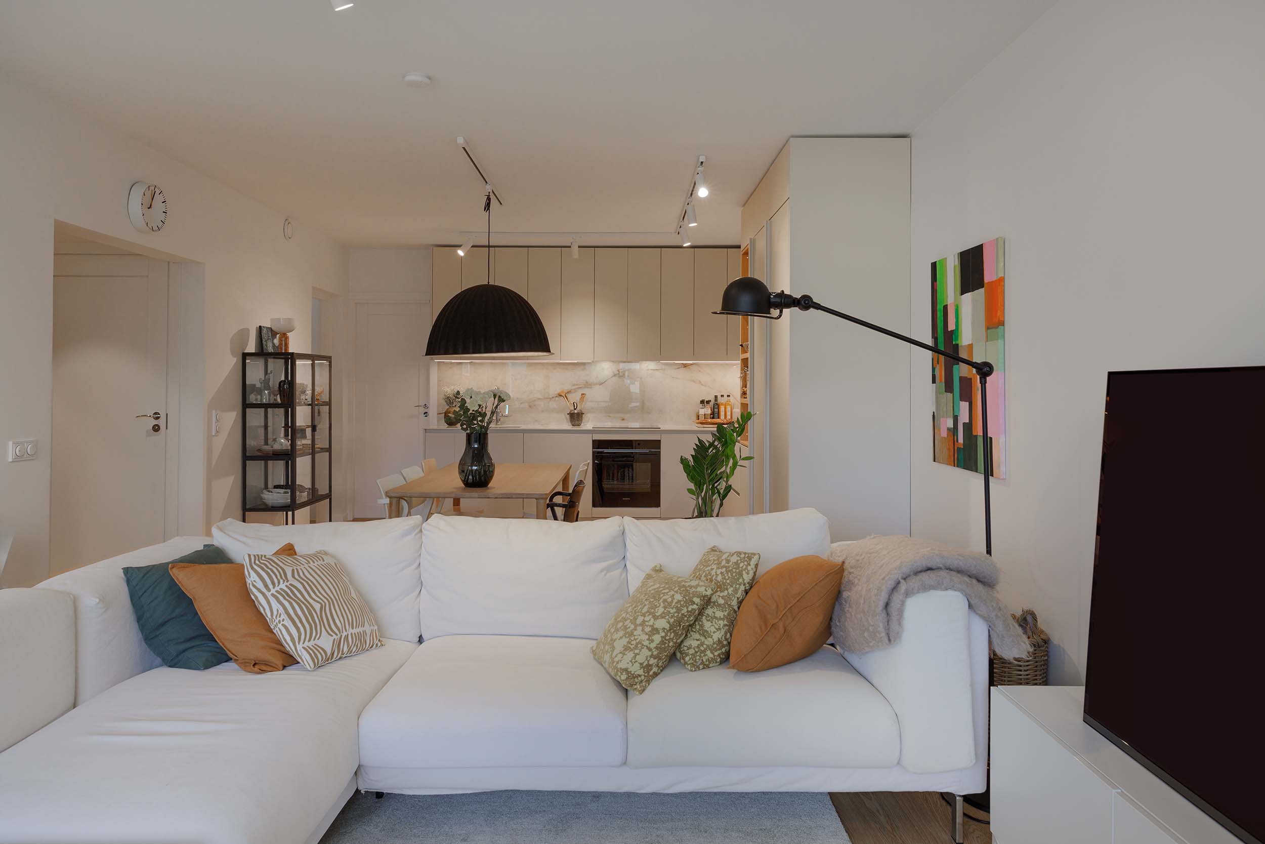 Modern living room with white sectional sofa adorned with decorative pillows and a blanket, facing a large flat-screen TV, with a dining table and kitchen in the background.