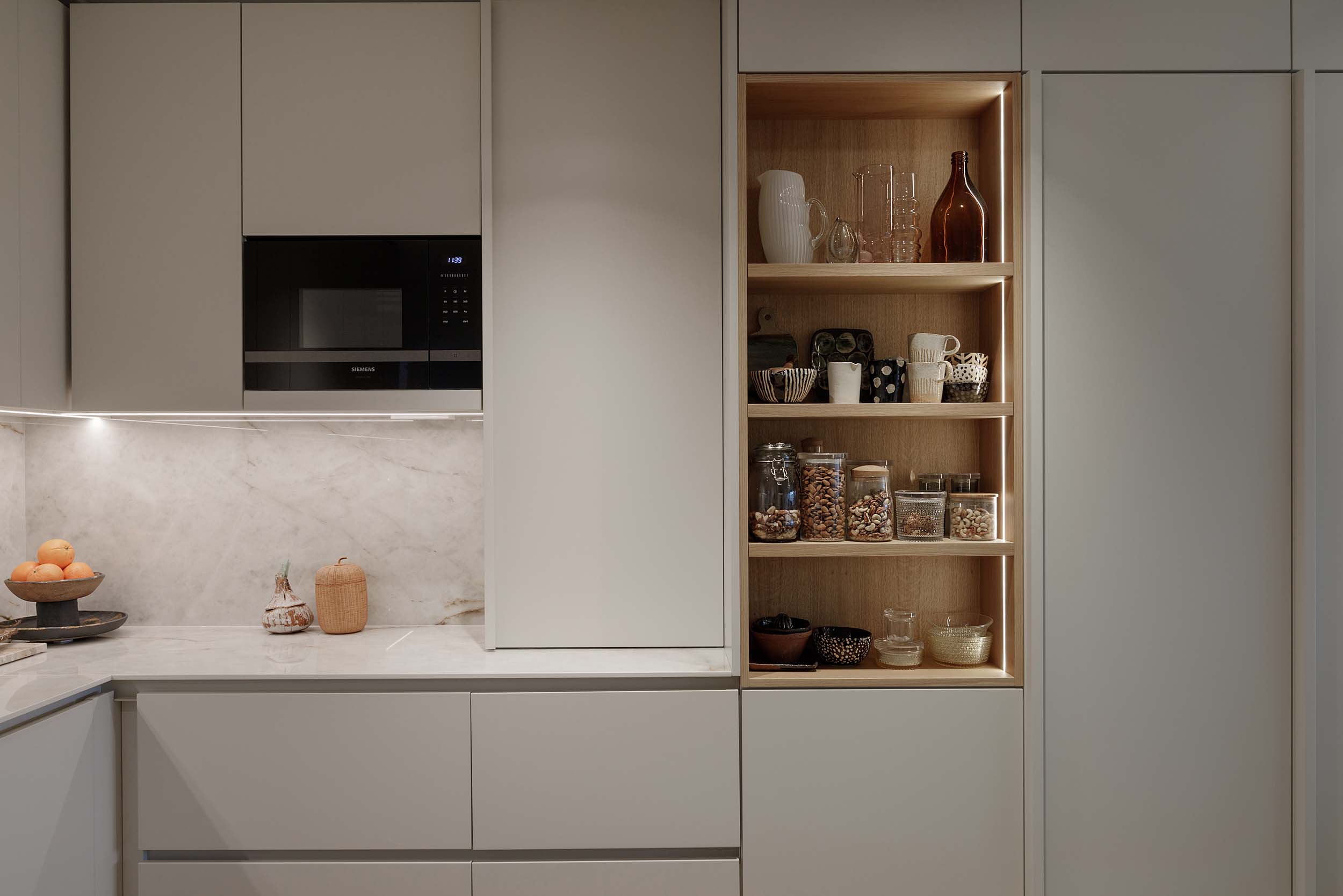 Modern kitchen with built-in black microwave, light gray cabinets, marble countertop, and wooden open shelves displaying glassware, cups, and jars of nuts.