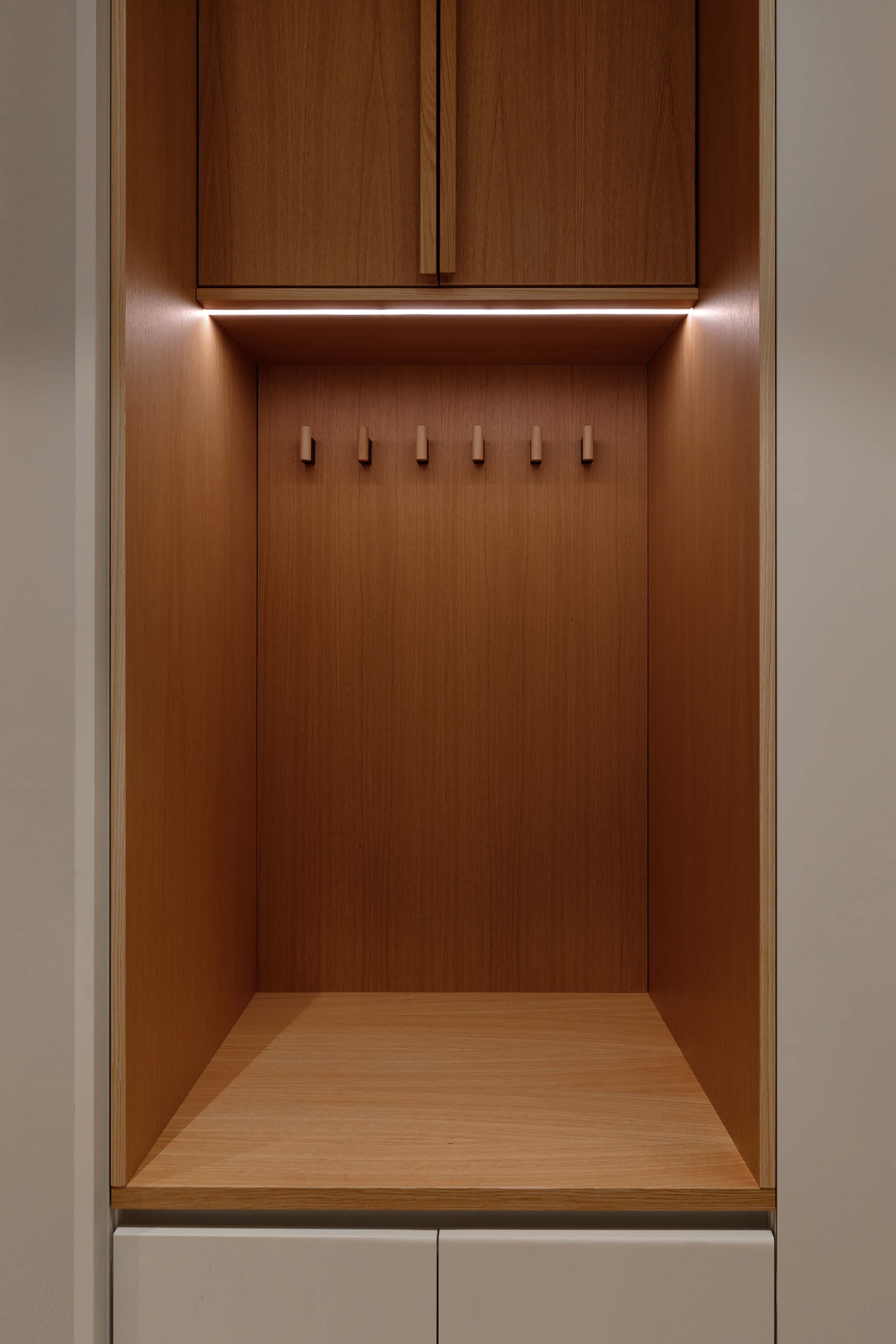Minimalist wooden built-in coat rack with seven hooks, upper cabinet, and soft lighting underneath.