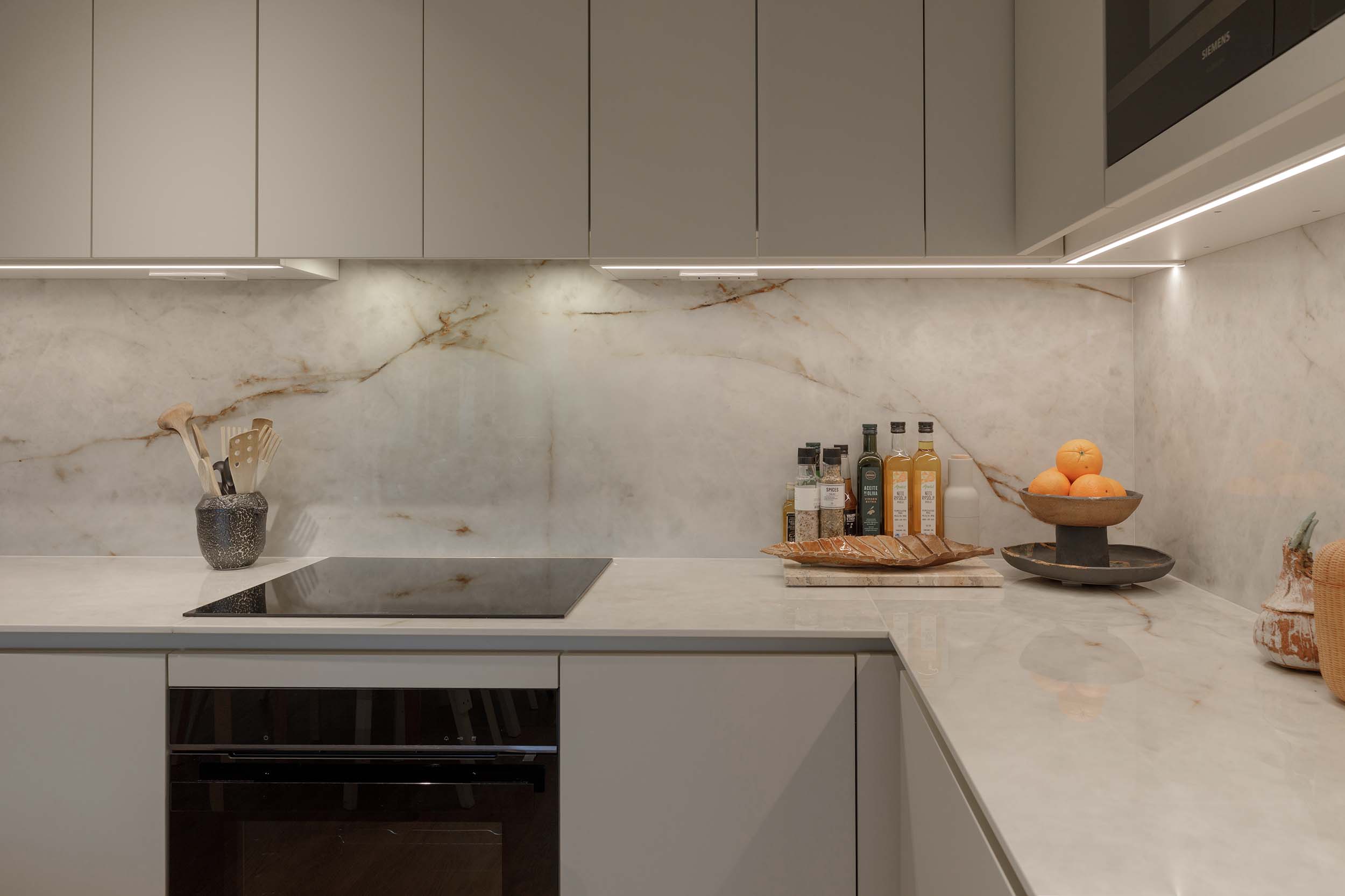 Modern kitchen countertop with built-in stovetop, kitchen utensils in a black container, bottles of oils and spices, a loaf of bread on a cutting board, and a bowl of oranges on a marble backsplash.