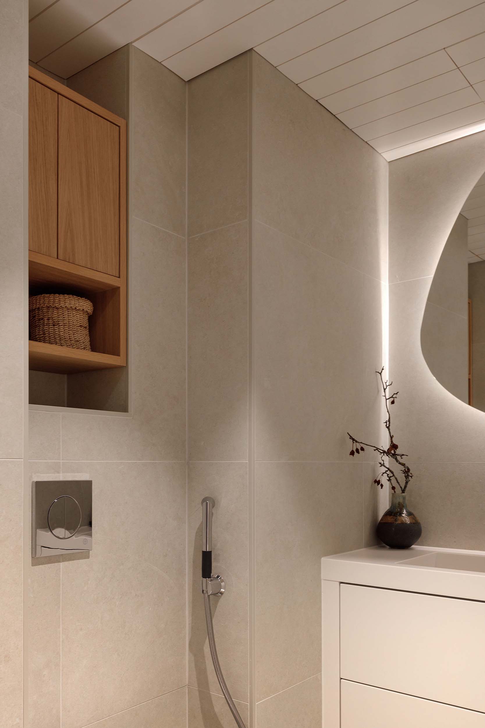 Minimalist bathroom corner with beige tiled walls, a white vanity with a vase holding dried branches, and a wall-mounted bidet spray.