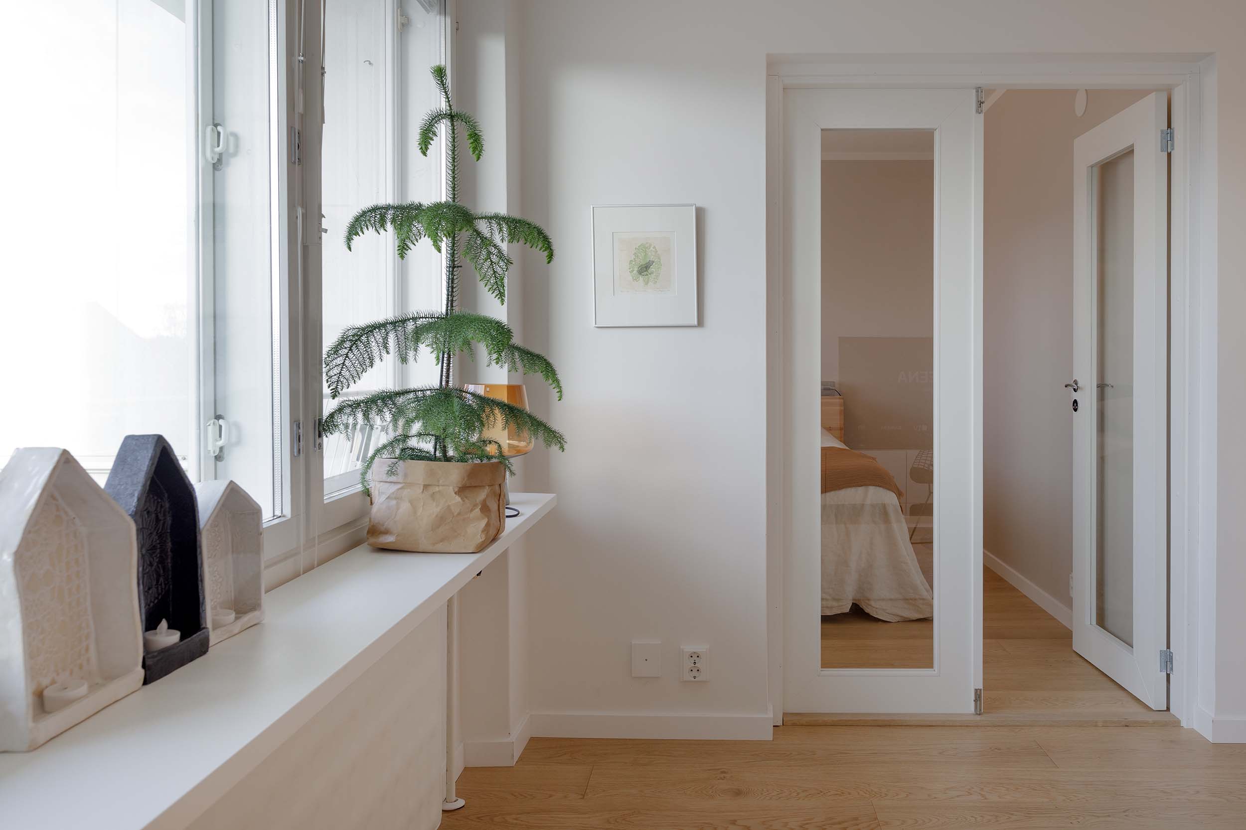 Minimalist indoor space with a small potted Norfolk Island pine on a white windowsill beside decorative house-shaped candle holders and a door with a glass panel showing a bedroom.
