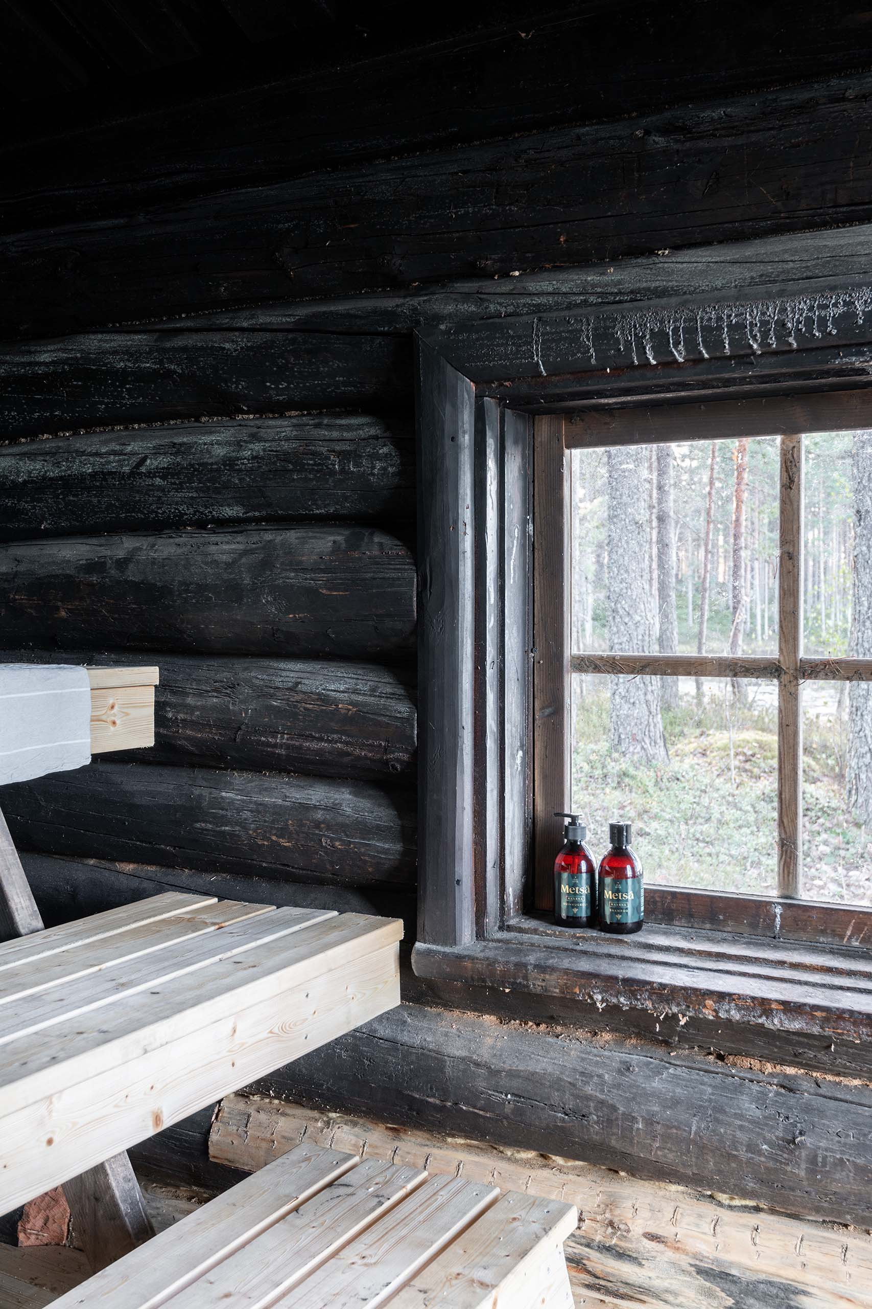 Interior of a rustic wooden sauna with light wood benches and a window showing forest trees outside with two red bottles on the windowsill.