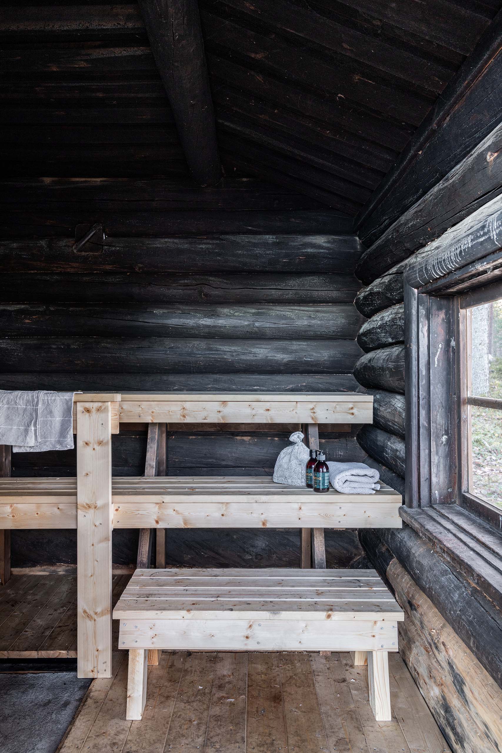 Interior view of a wooden sauna with light-colored benches, a sauna hat, towel, and bottles placed on the upper bench next to a window.