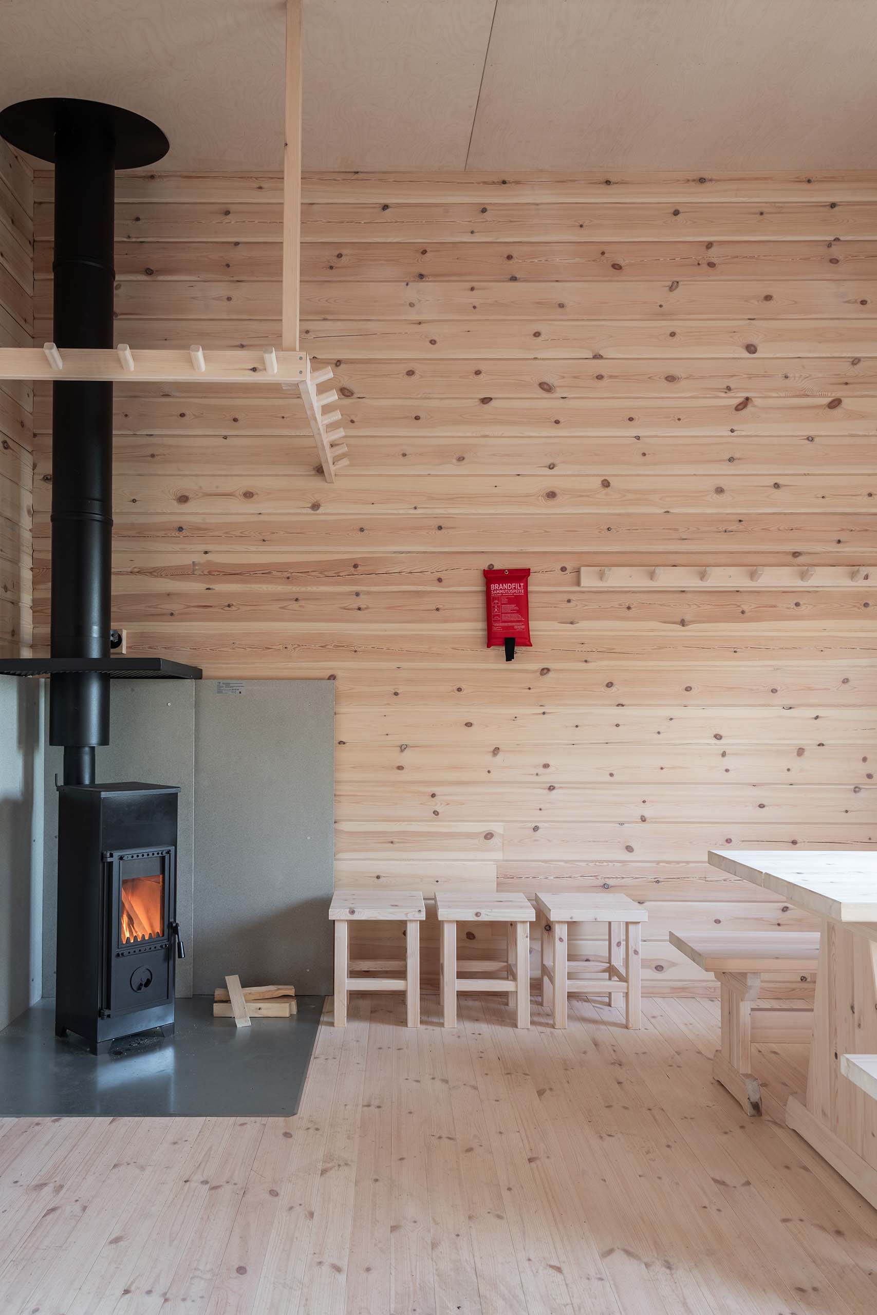Interior of a wooden room with a lit black wood stove, three wooden stools, a wooden table, and a red fire blanket on the wall.