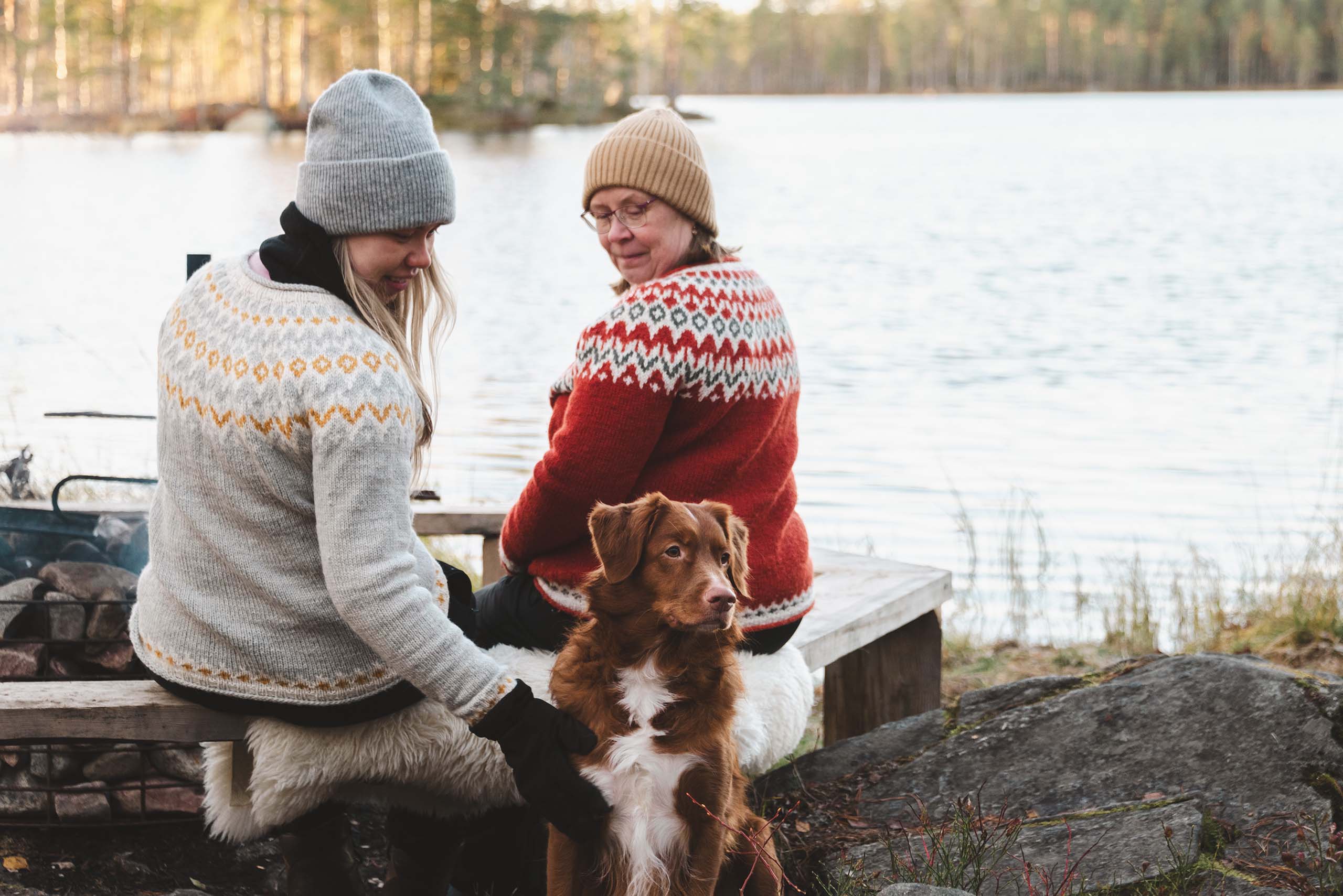 Two women in knitted sweaters and hats sitting by a lake with a brown and white dog in the foreground.
