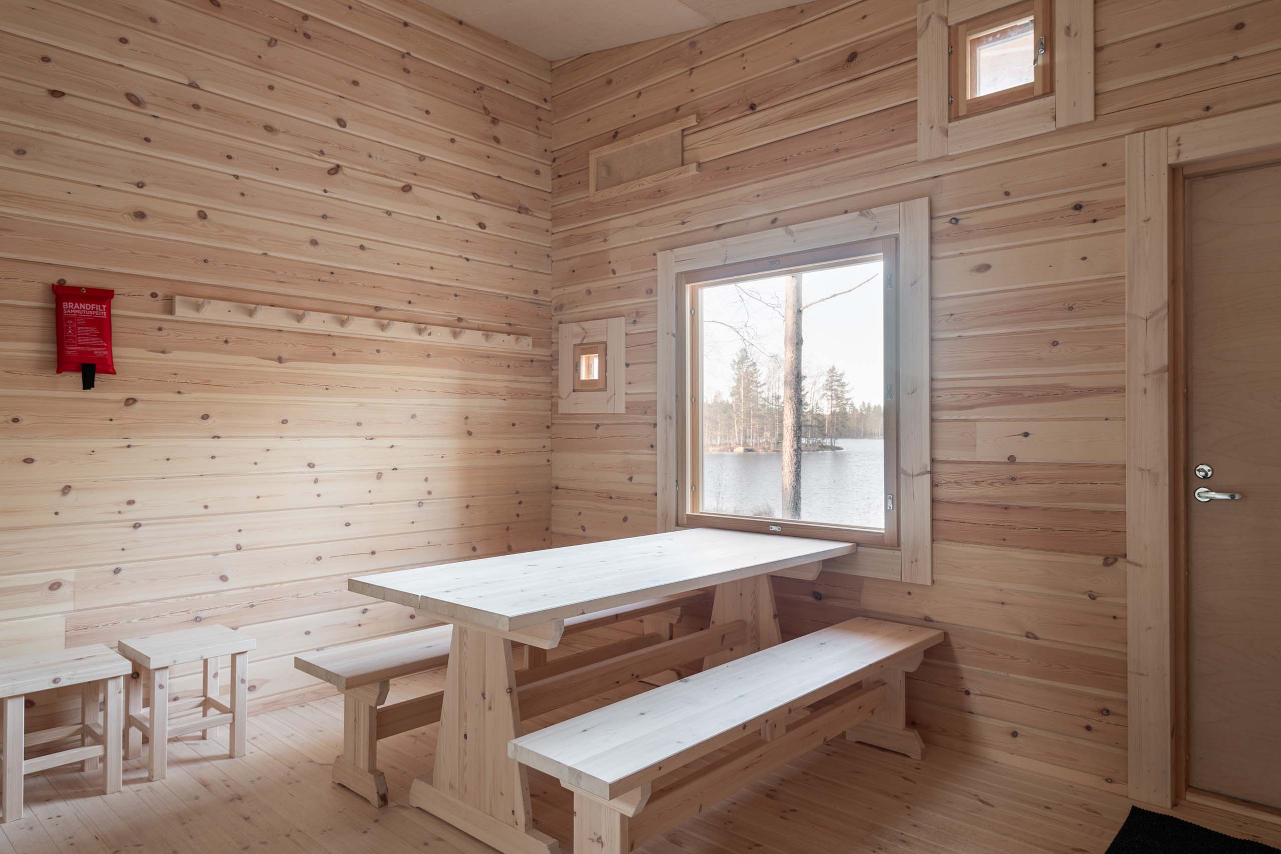 Minimalist wooden room with a light wood table, benches, stools, and a window showing a lake view with trees.