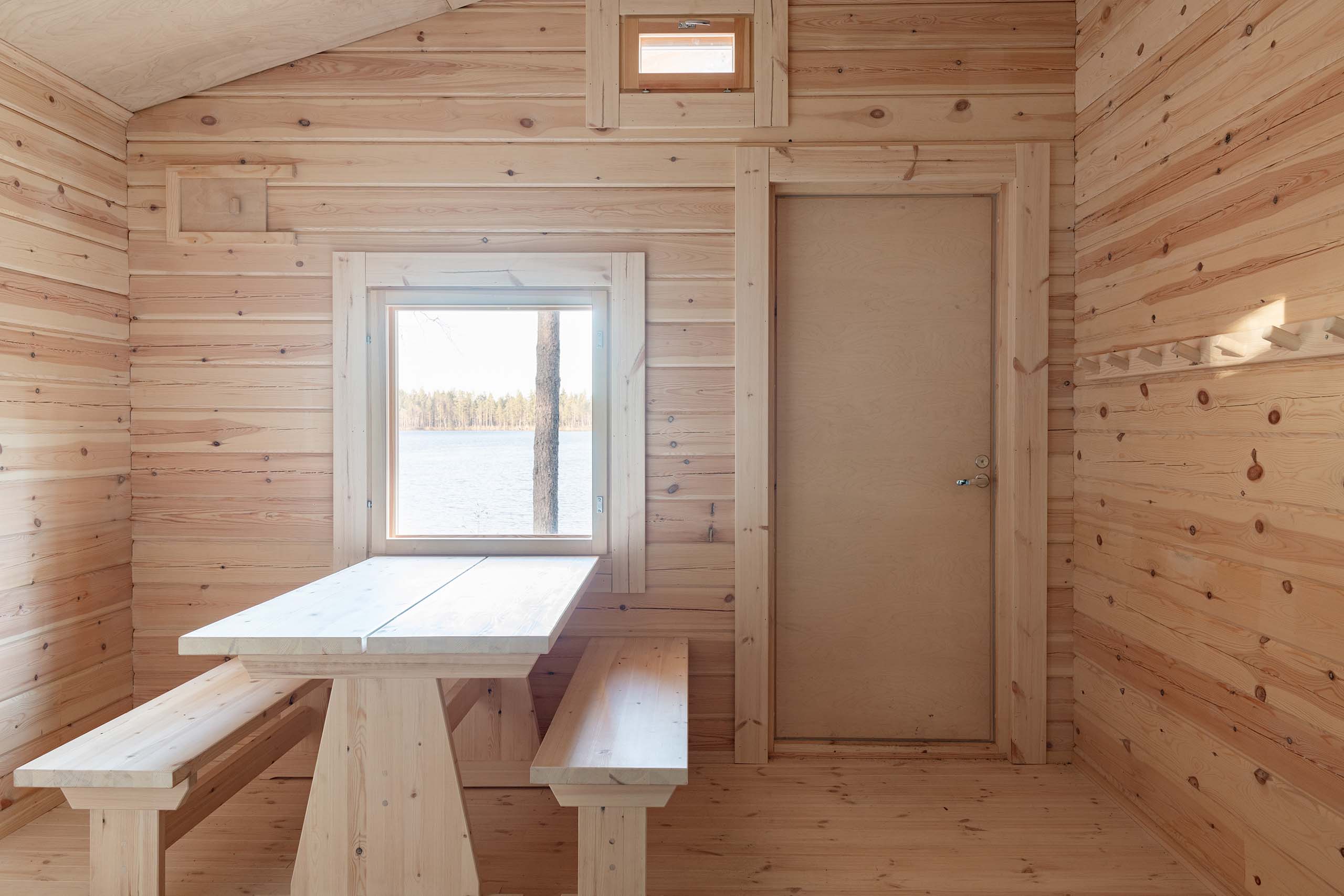 Interior of a wooden cabin room with a light wood table and benches, a closed door, and a window showing a lake and trees outside.