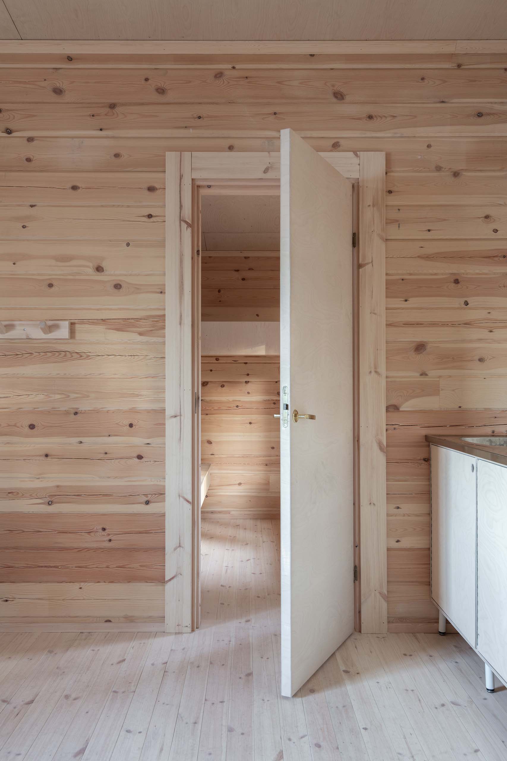 Interior view of a wooden room with light wood walls and floor, featuring a partially open plain white door and a white cabinet to the right.