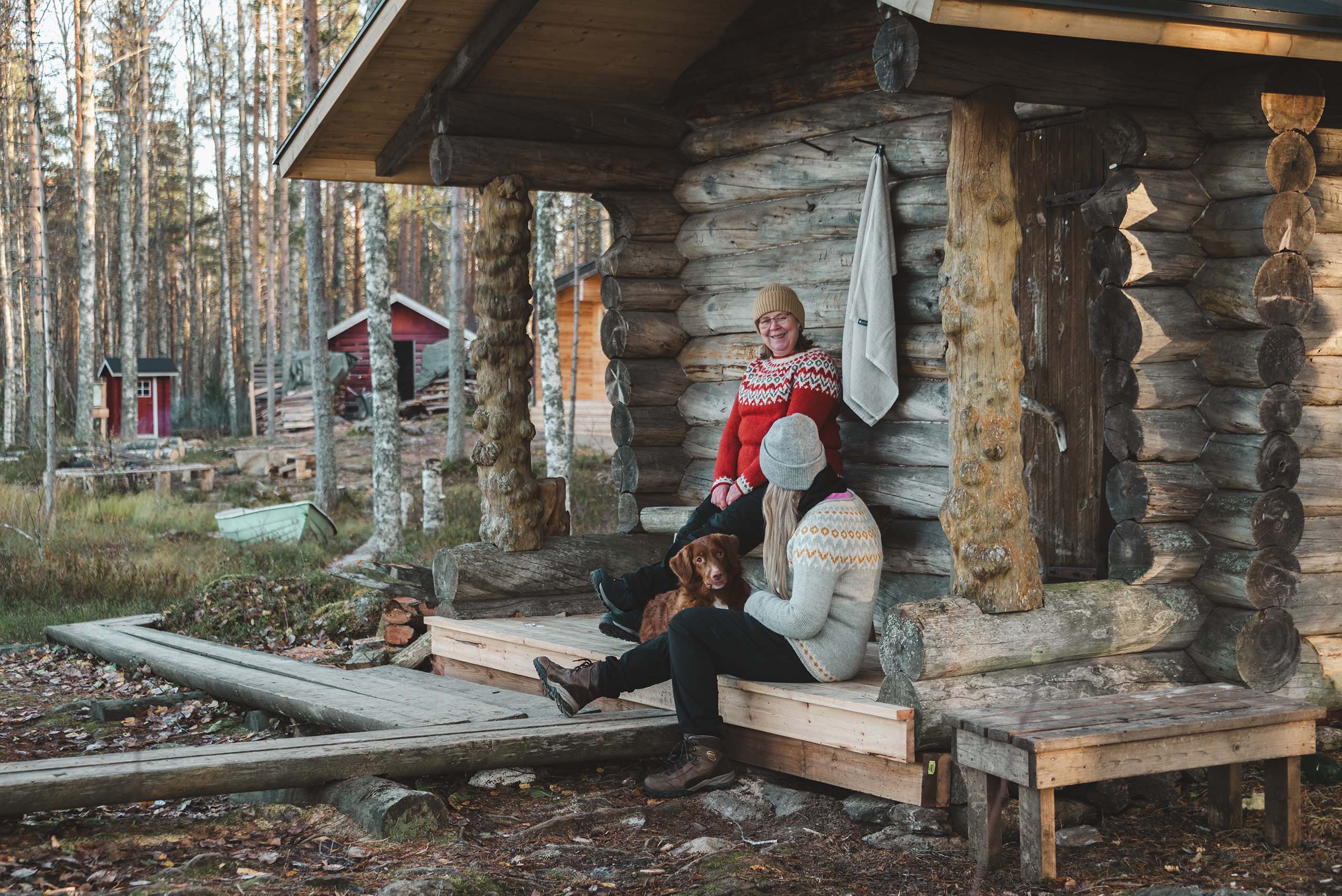 Two people in knitted sweaters sitting on the porch of a rustic log cabin, with a brown dog between them and a forest in the background.