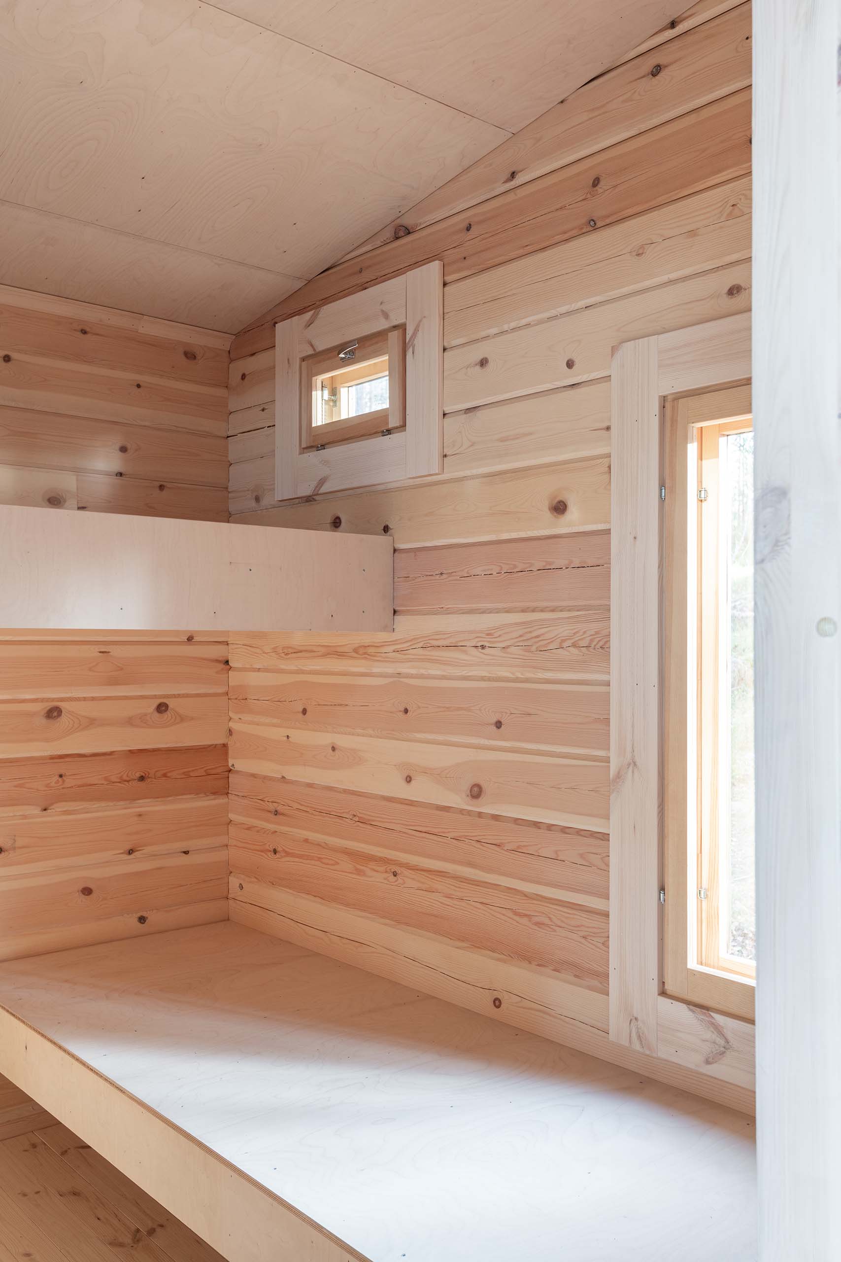 Interior of a wooden cabin room with unfinished bunk beds and natural light coming through two small windows.