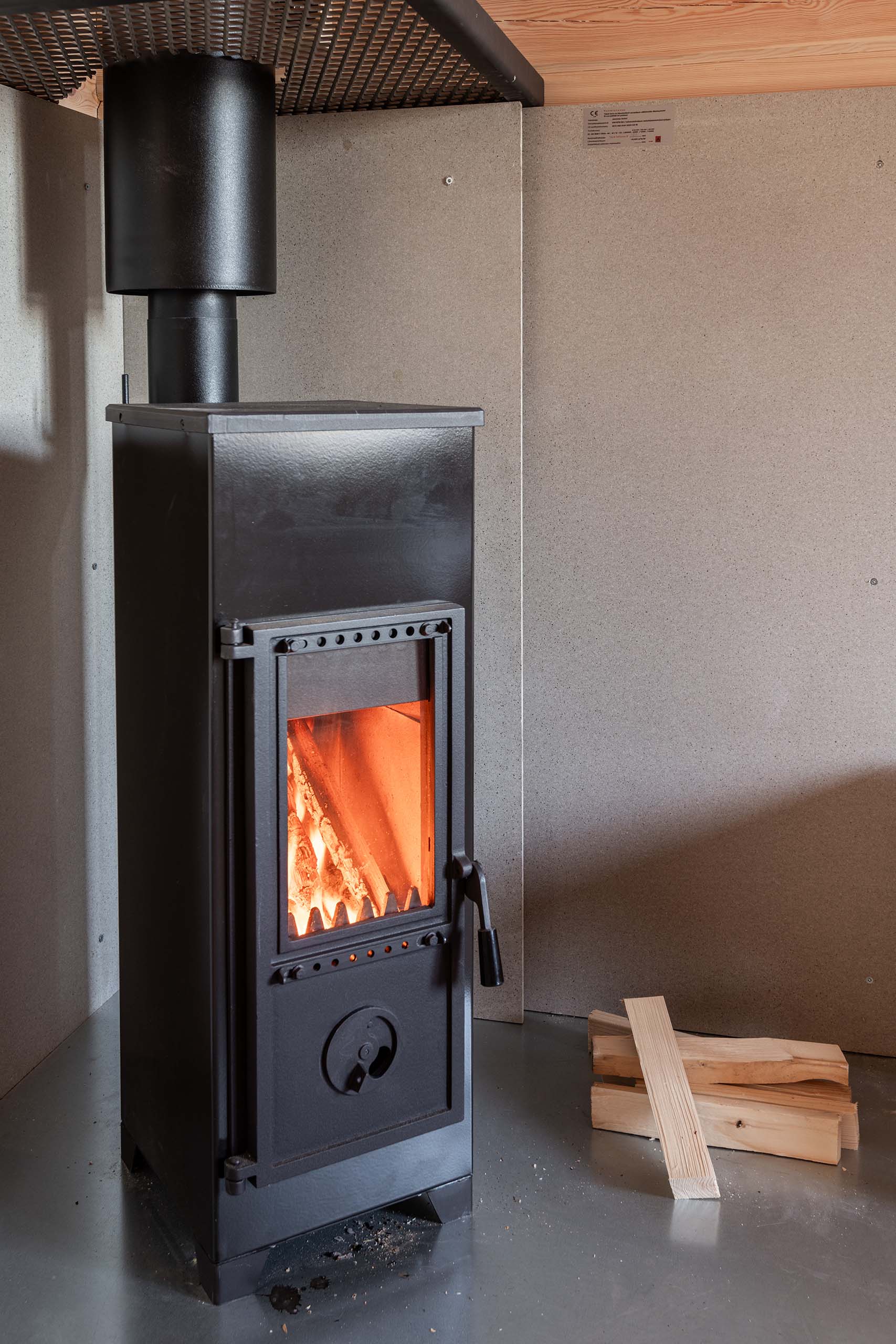 Black wood-burning stove with fire visible through glass door next to a small stack of firewood on a gray floor with beige walls.