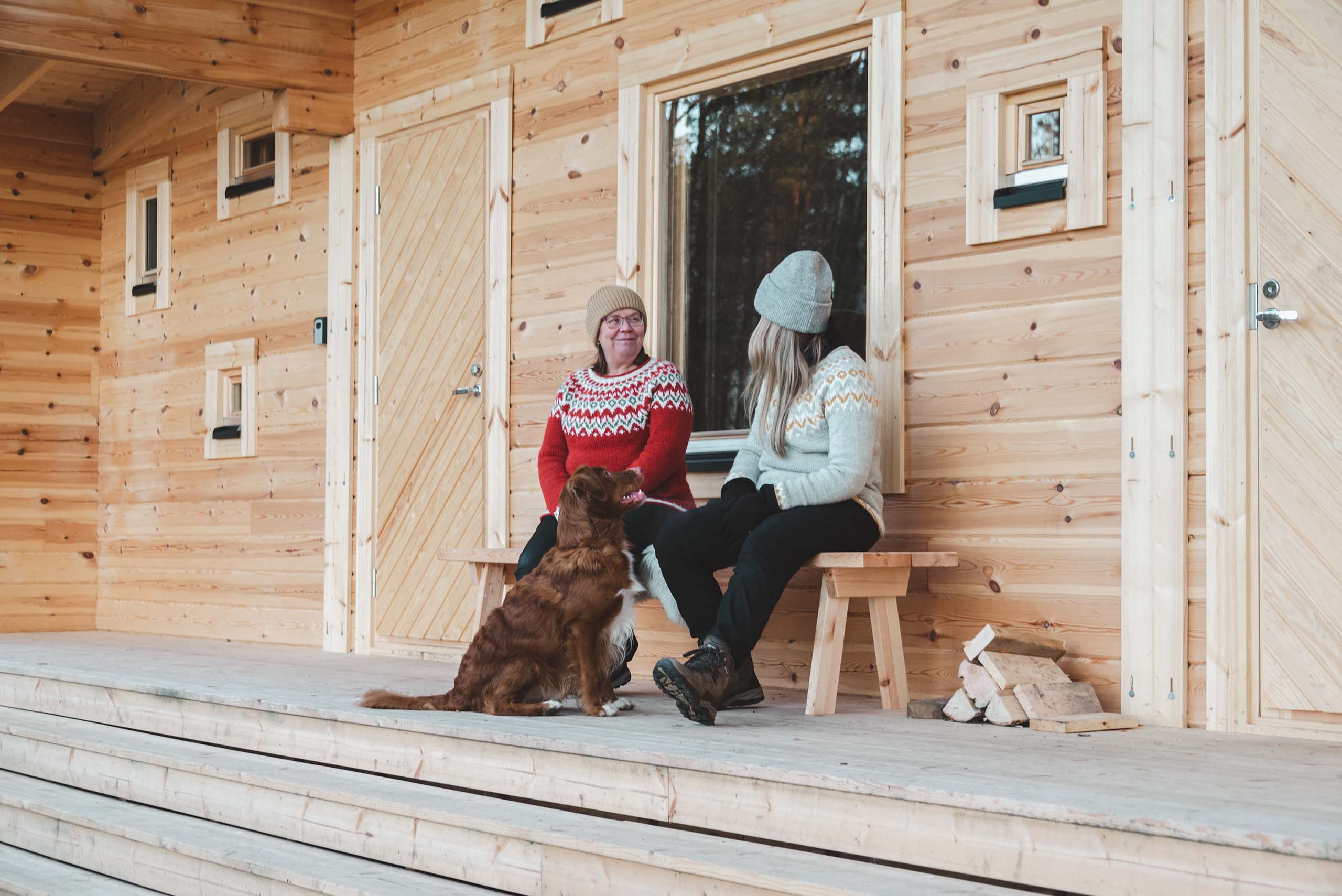 Two women in knitted sweaters and beanies sitting on a wooden bench on a porch with a brown dog sitting on the floor in front of them.