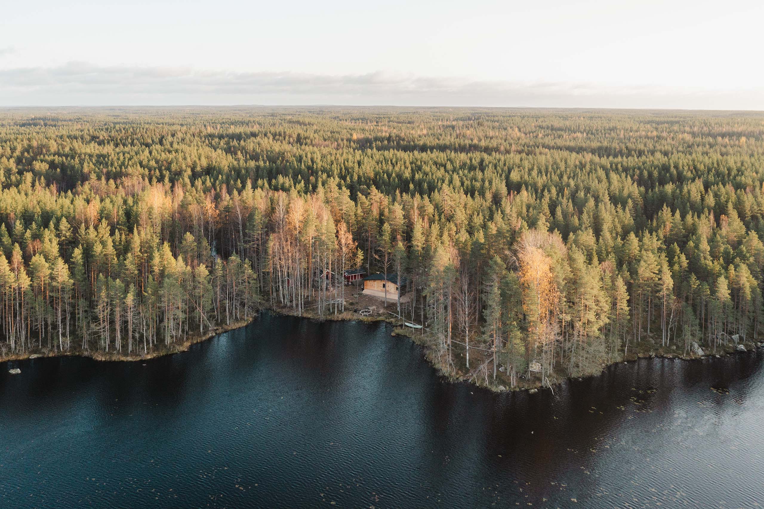 Aerial view of a cabin surrounded by autumn-colored trees beside a dark blue lake reflecting the forest.