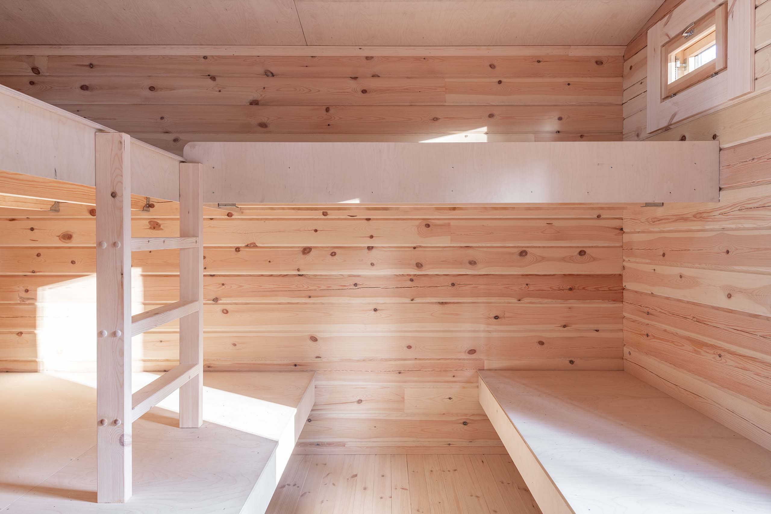 Interior of a wooden room with light natural wood bunk beds and walls, illuminated by sunlight through a small window.