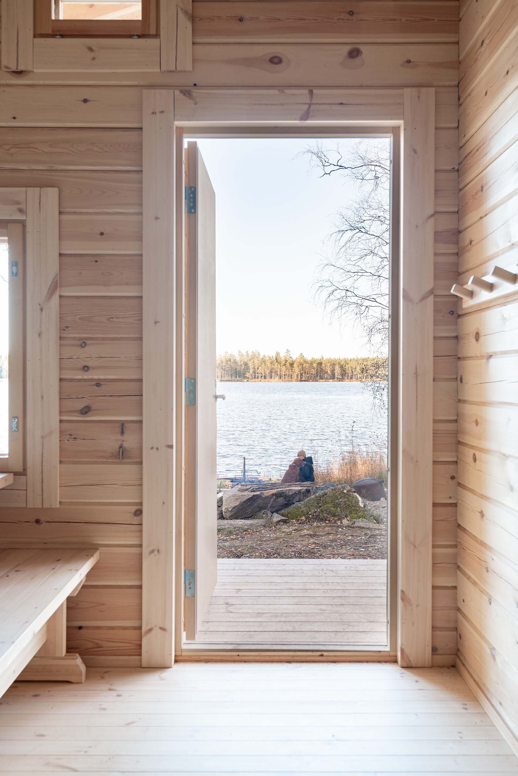 View through an open door of a wooden cabin showing a person sitting with a dog by a lakeside surrounded by trees.