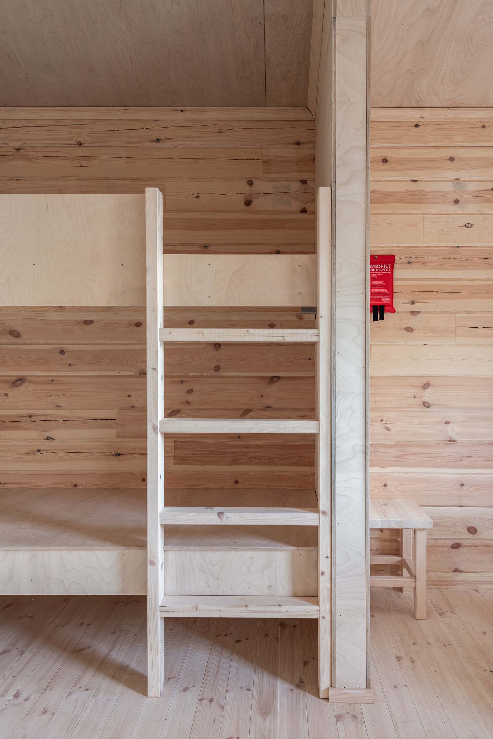 Minimalist wooden bunk bed ladder against light wood-paneled wall with small wooden bench and red fire safety bag.