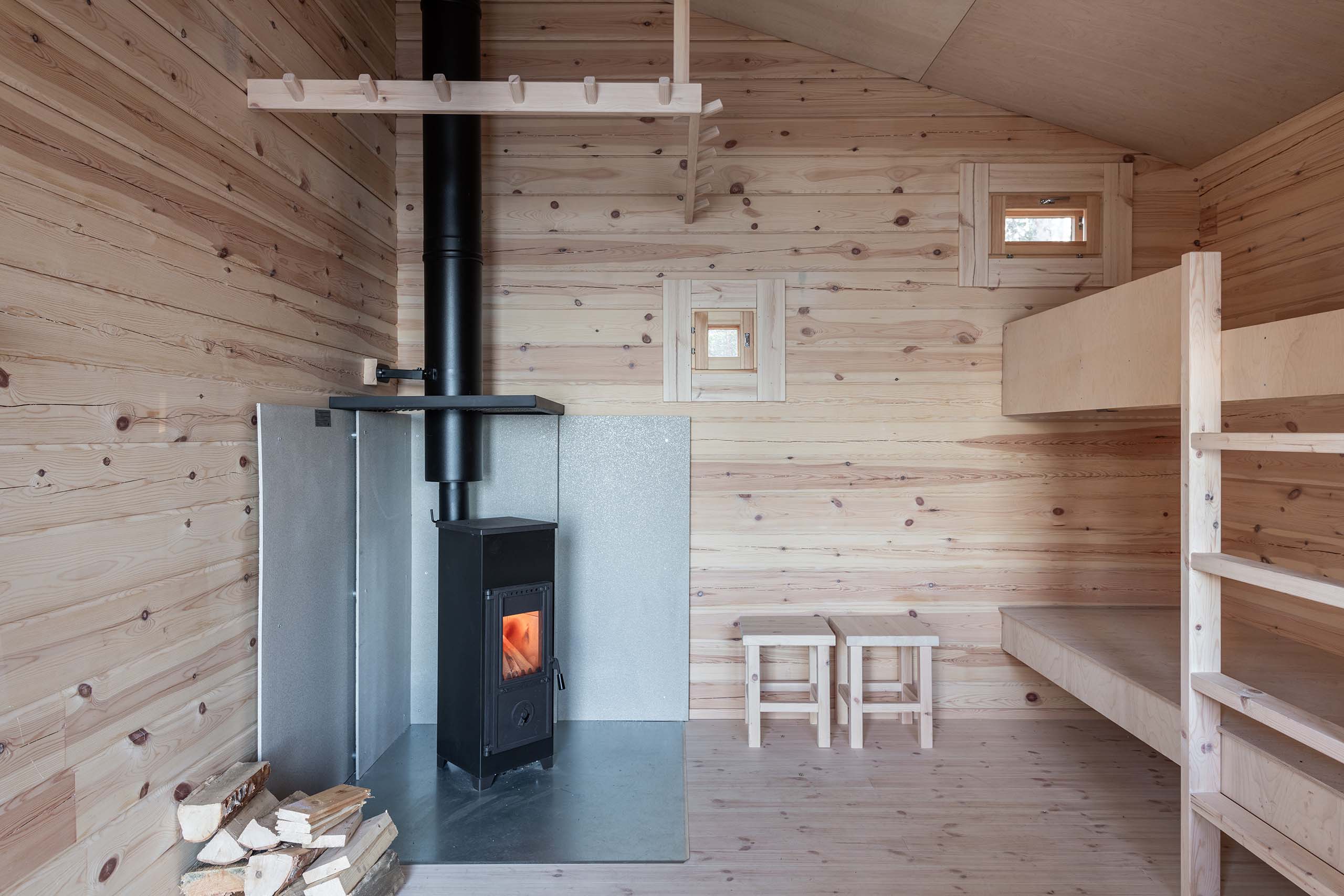 Minimalist wooden cabin interior with a small black wood-burning stove, stacked firewood, two wooden stools, and a built-in bunk bed.