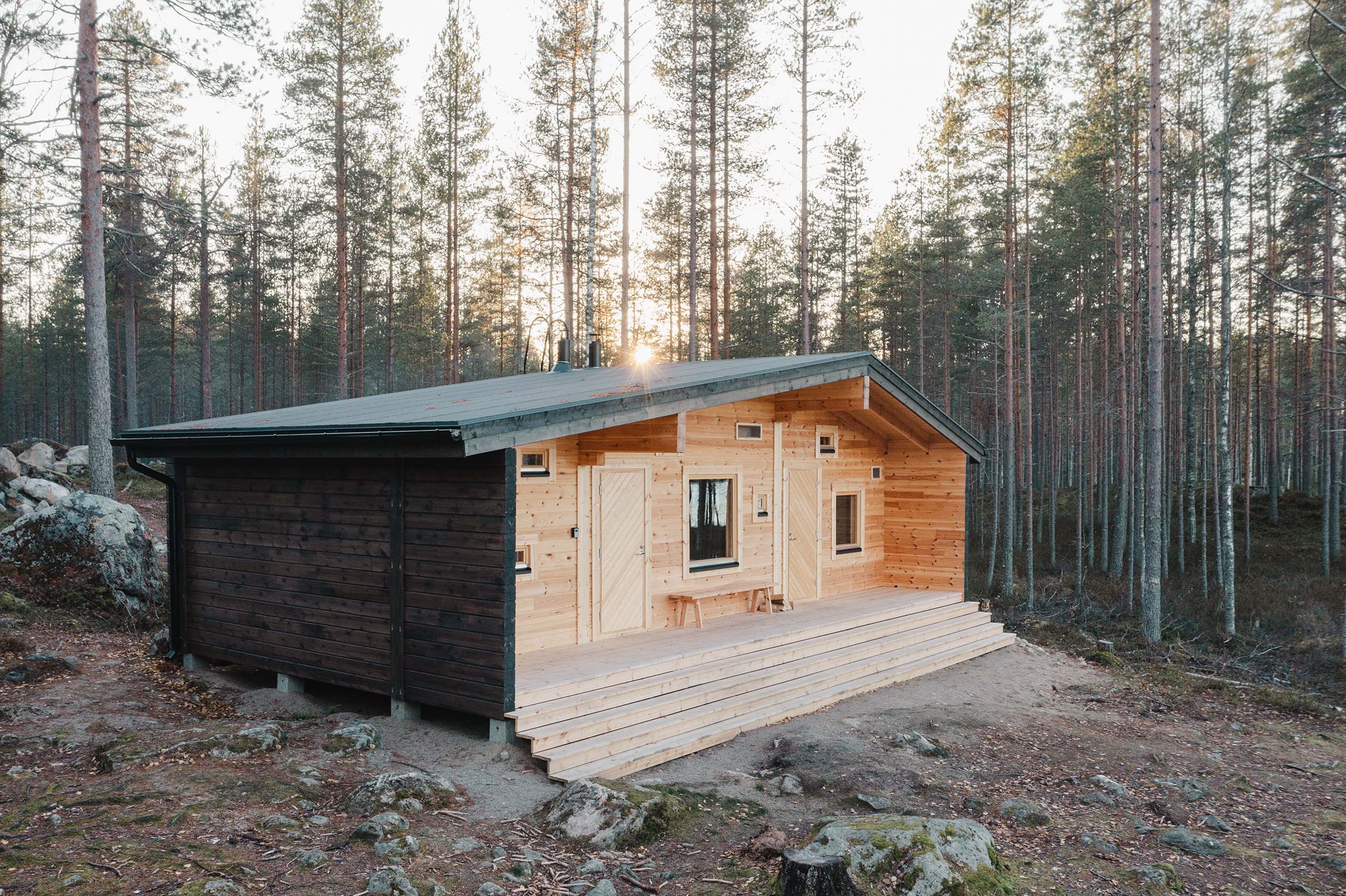 Wooden cabin with light and dark wood panels in a forest with tall pine trees during sunset.