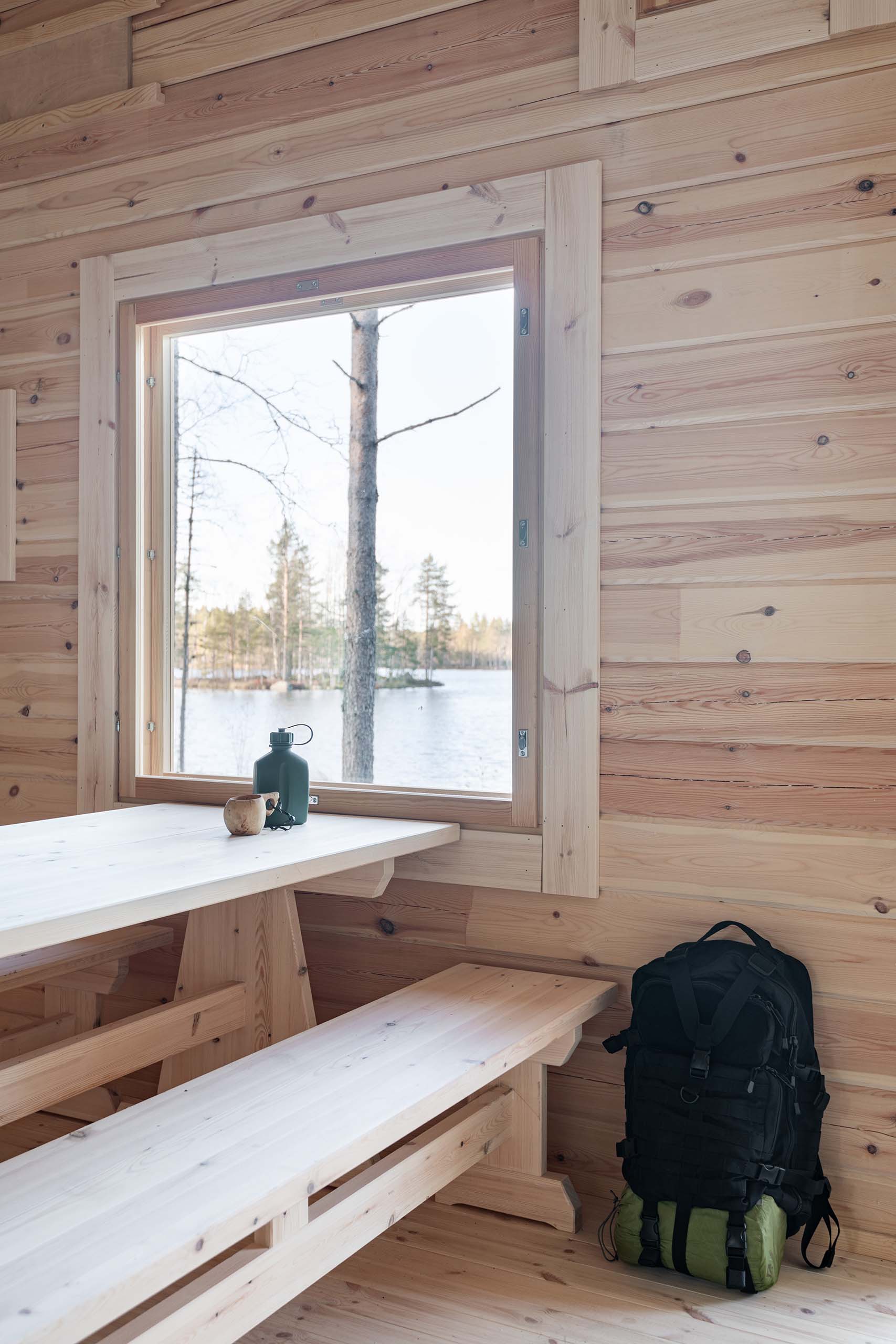 Wood-paneled cabin interior with a picnic table and bench, a window showing a lake and trees outside, a black backpack on the floor, and a green canteen and wooden cup on the table.