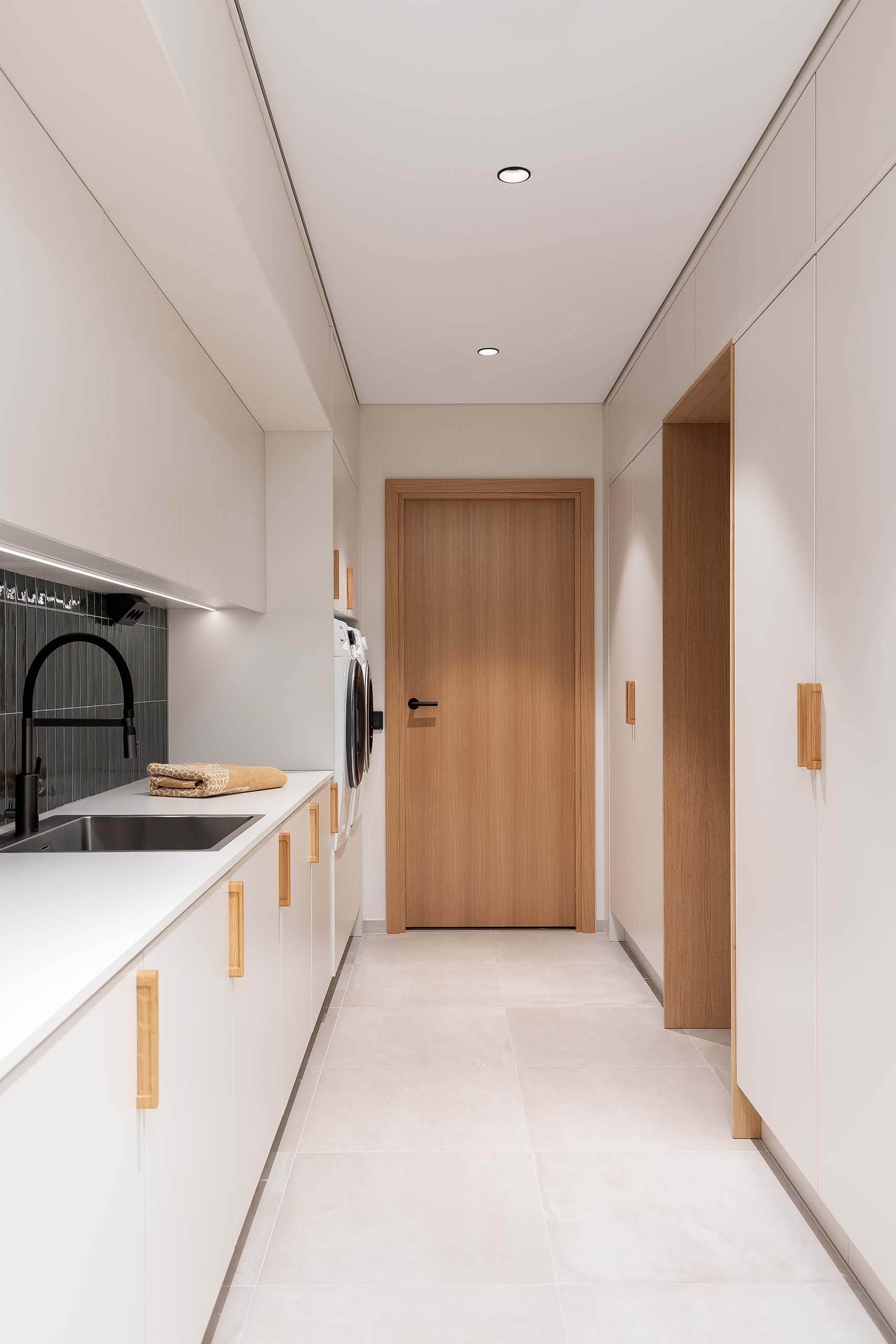 Modern laundry room with white cabinets, wooden handles, black faucet, washer and dryer, and a wooden door at the end.