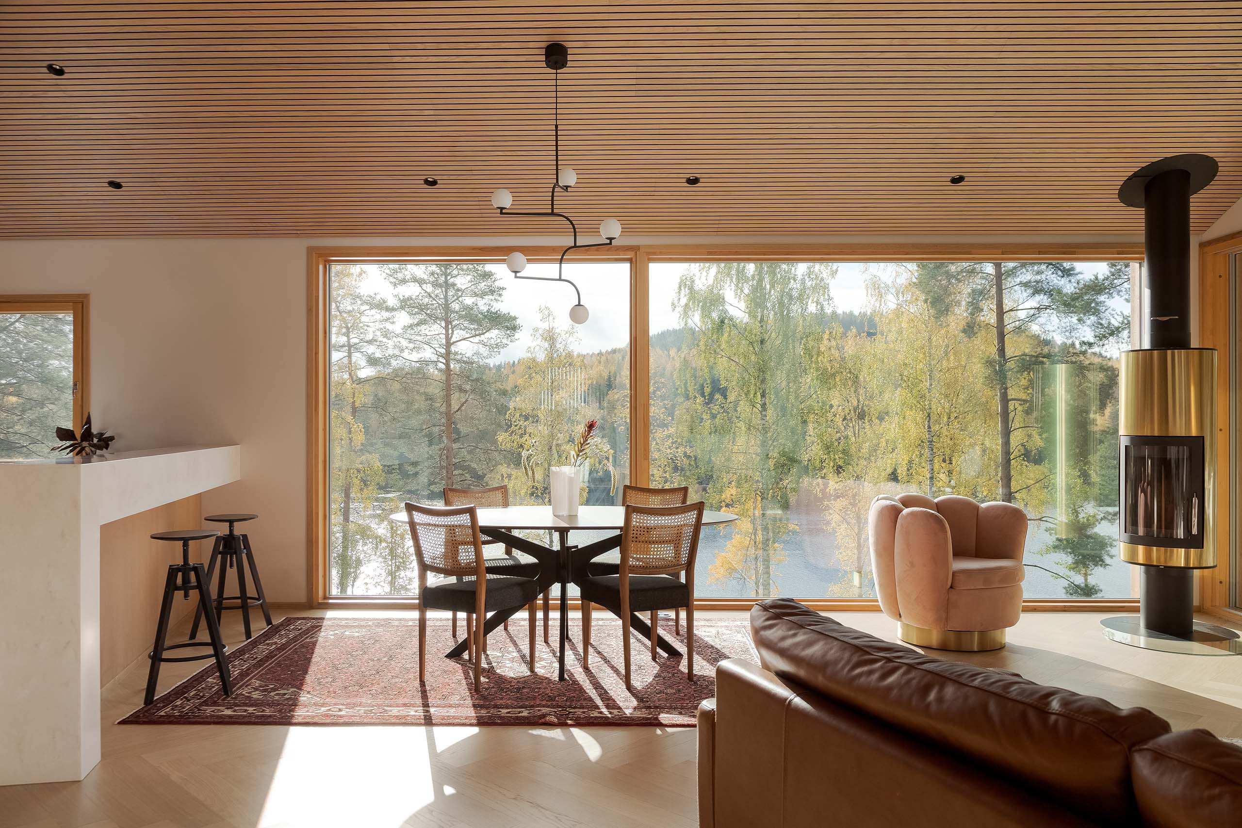Modern living room with large windows overlooking trees and water, featuring a round dining table with four chairs on a rug, a pink swivel chair, a leather sofa, and a wall-mounted fireplace.