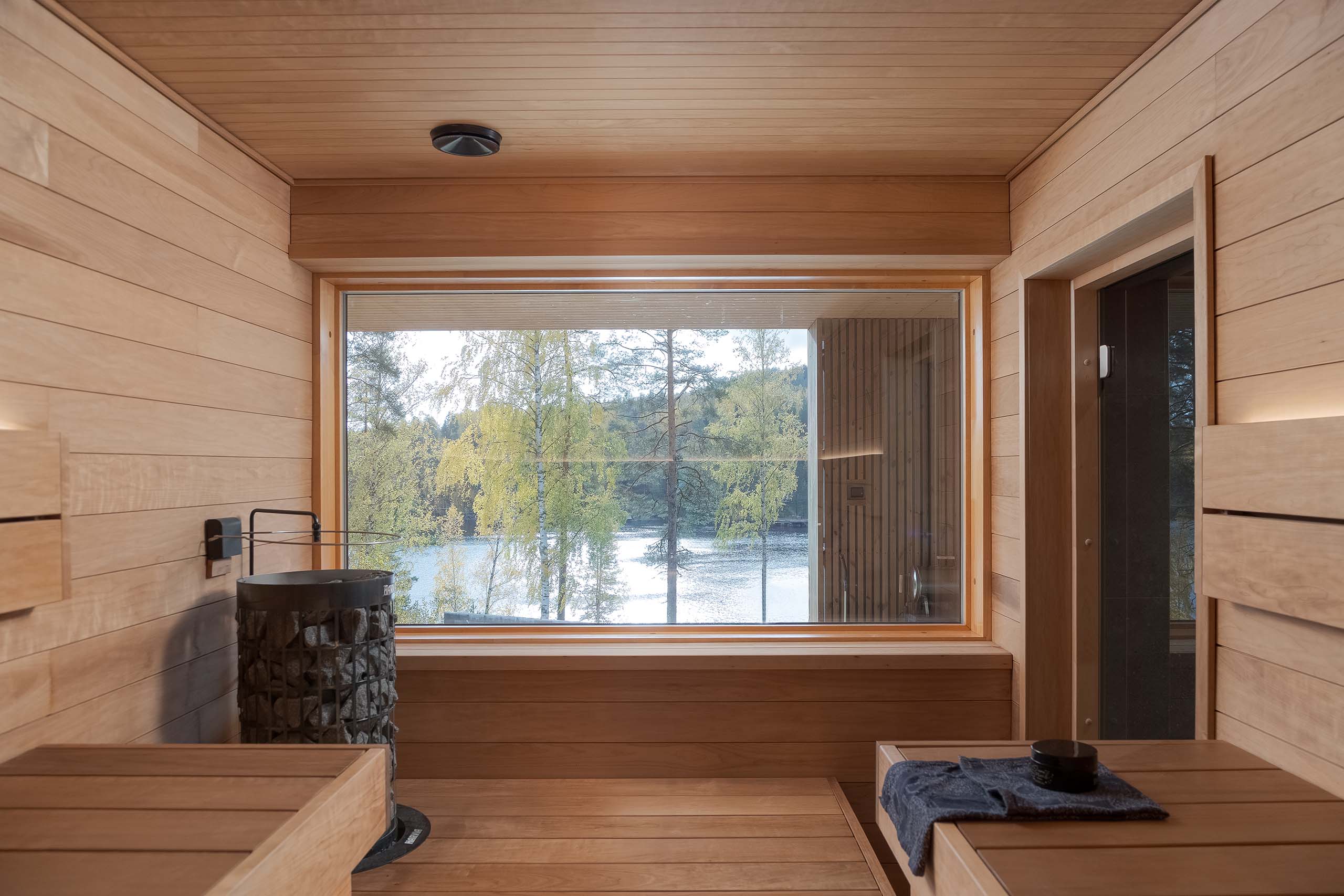 Interior of a wooden sauna with a large window overlooking a lake and trees outside.