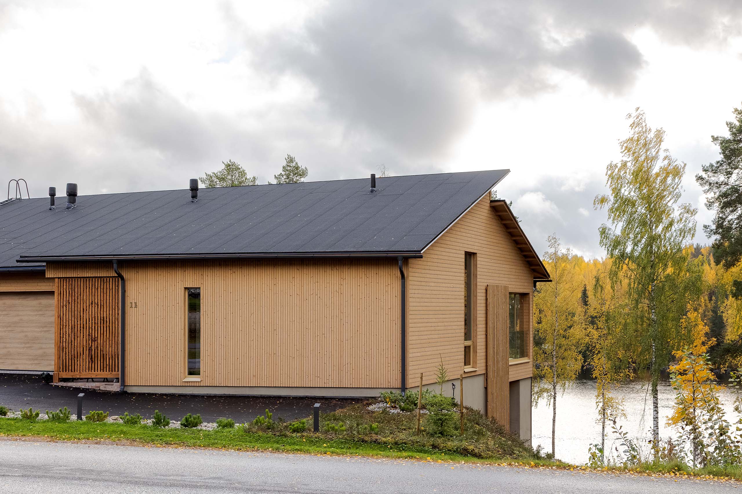 Modern wooden house with a dark sloped roof beside a road, surrounded by autumn trees and overlooking a lake.