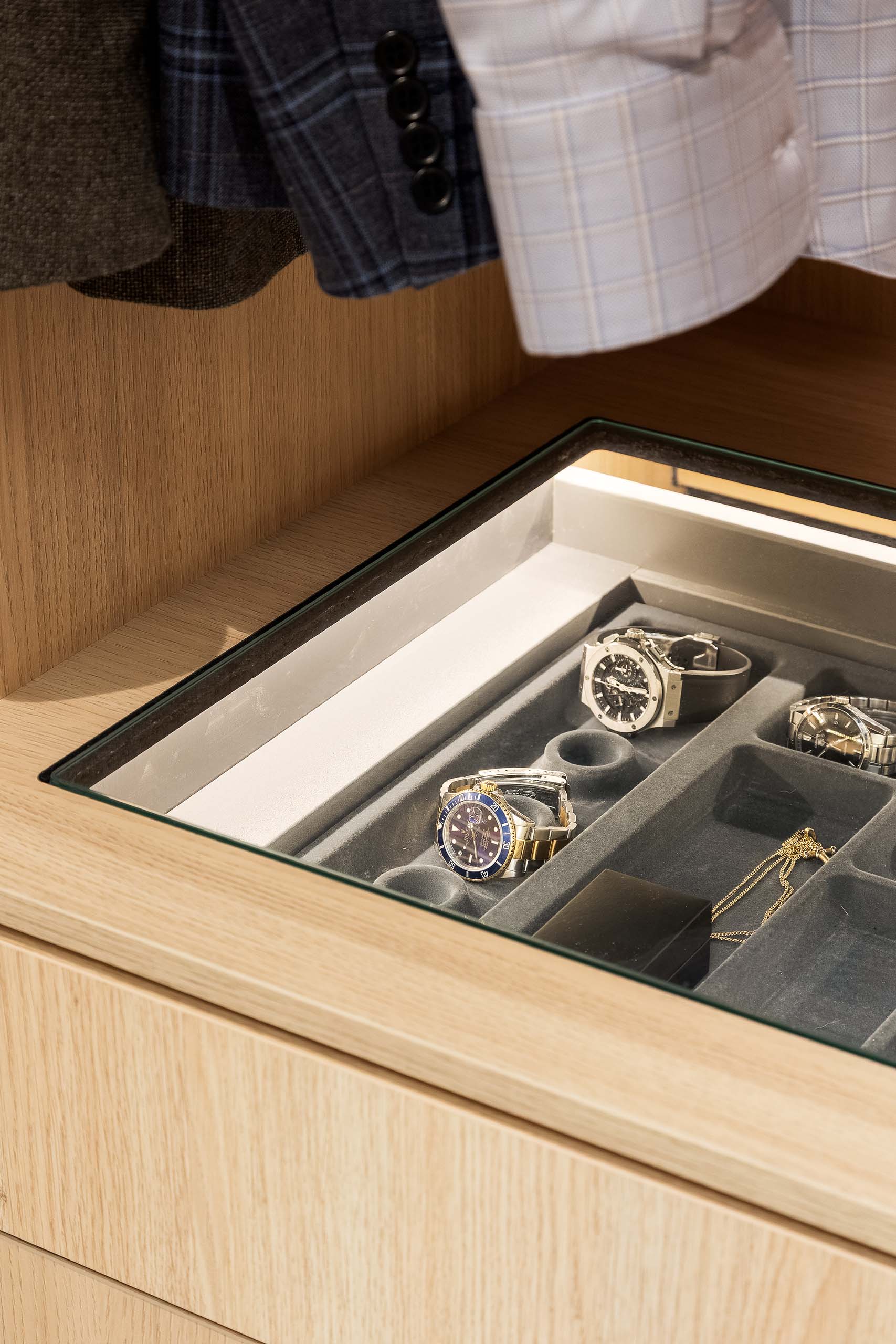 A wooden drawer with a glass top displaying multiple wristwatches and a gold chain inside a gray velvet organizer, with hanging clothes above.