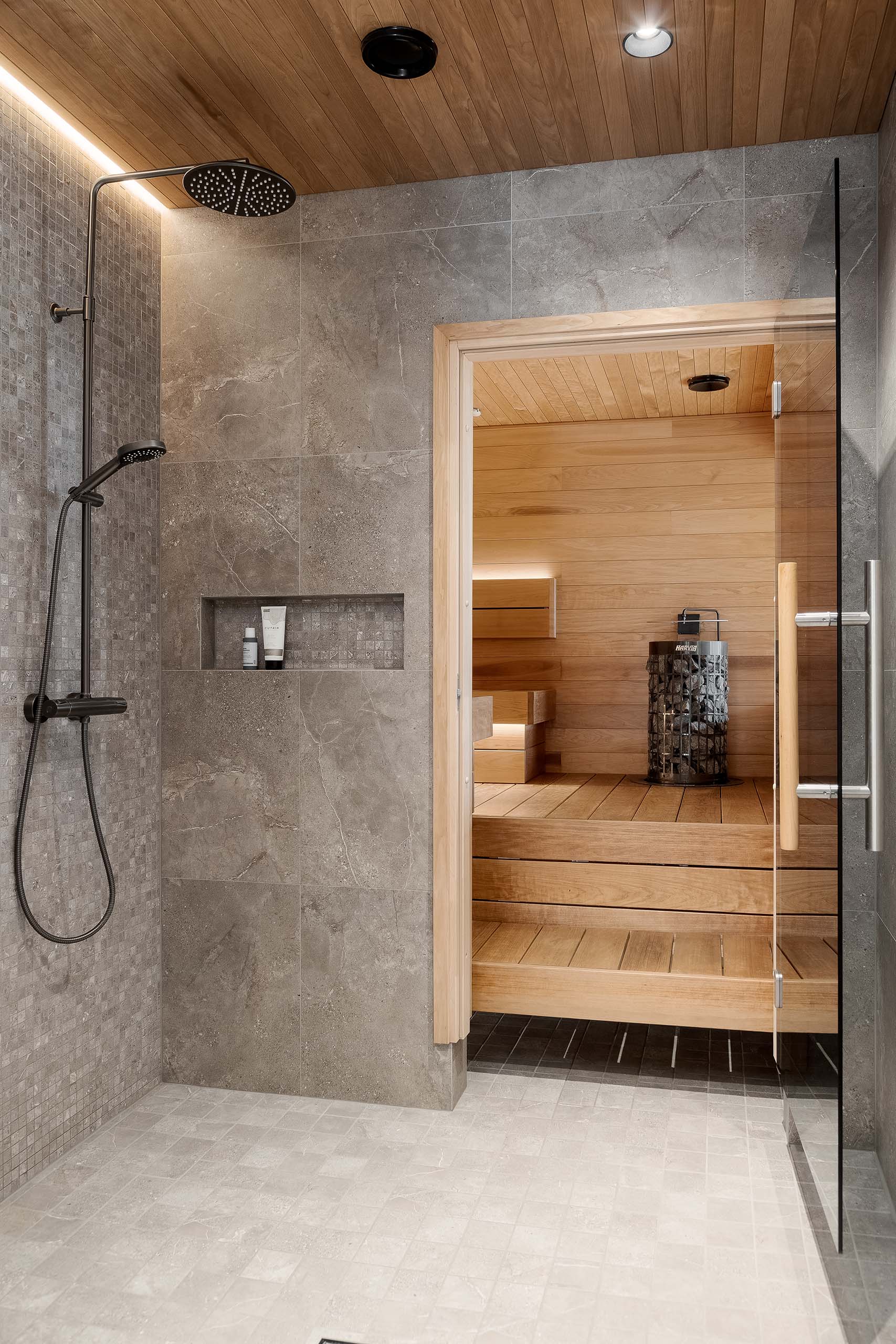 Modern shower area with dark rain showerhead and handheld shower next to a wooden sauna with glass door.