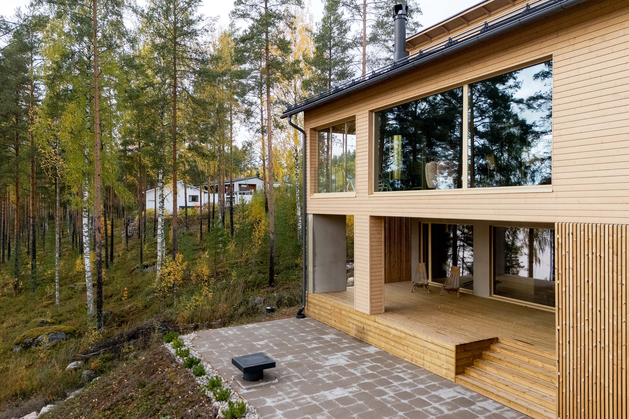 Modern wooden house with large glass windows and a patio, surrounded by a forest with tall trees during autumn.