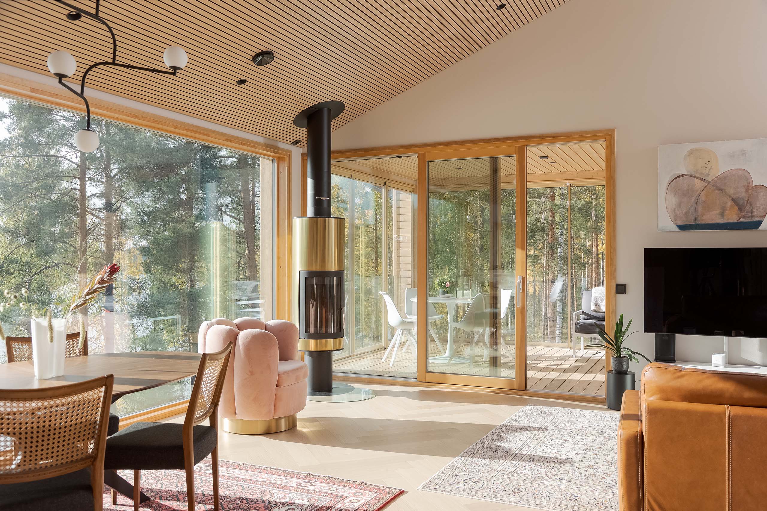 Modern living room with large glass windows overlooking trees, featuring a round fireplace, pink velvet chair, wooden dining table, and sliding glass doors to a deck with white chairs.