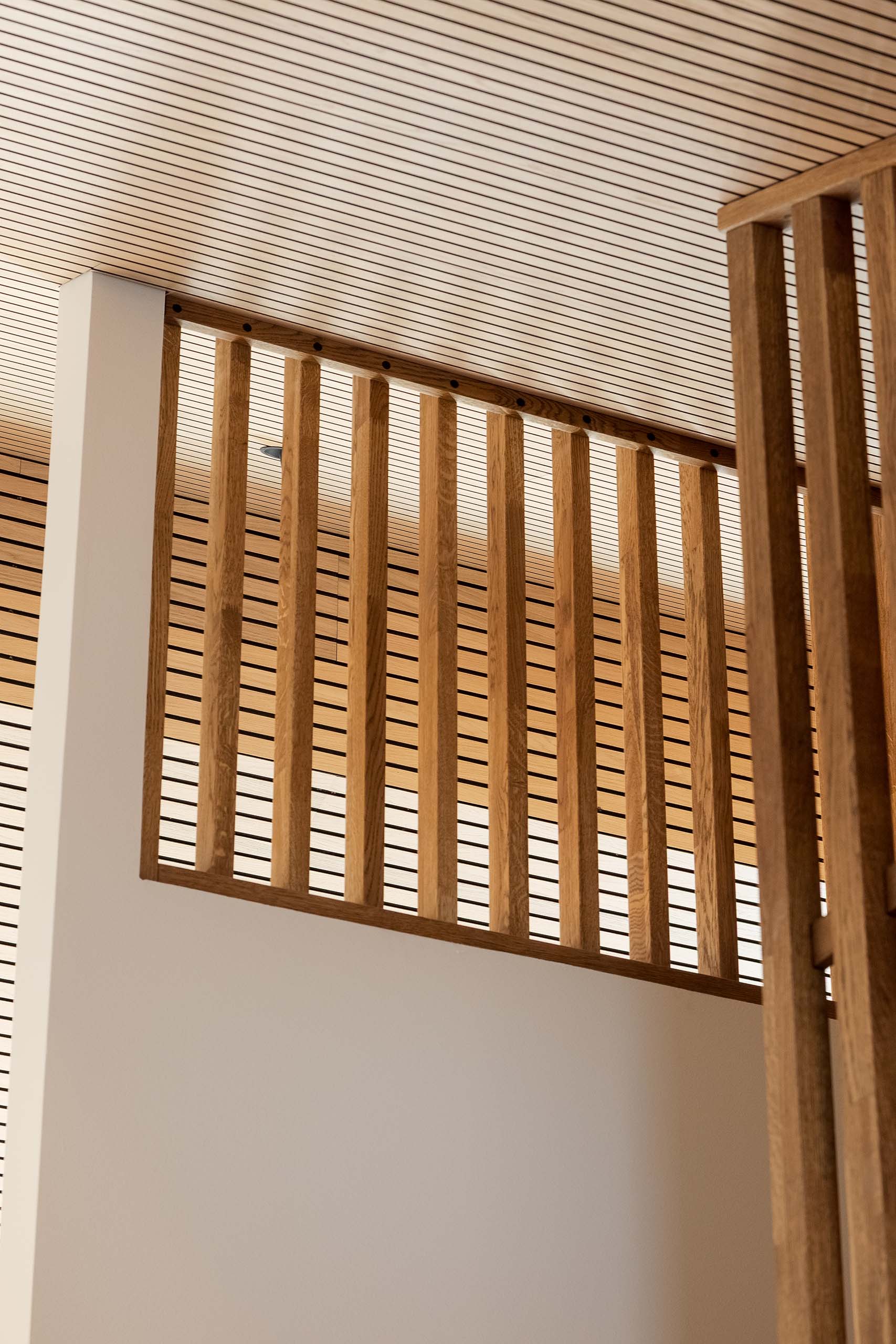 Wooden vertical slats of a stair railing against a ceiling with parallel black lines on light wood panels.