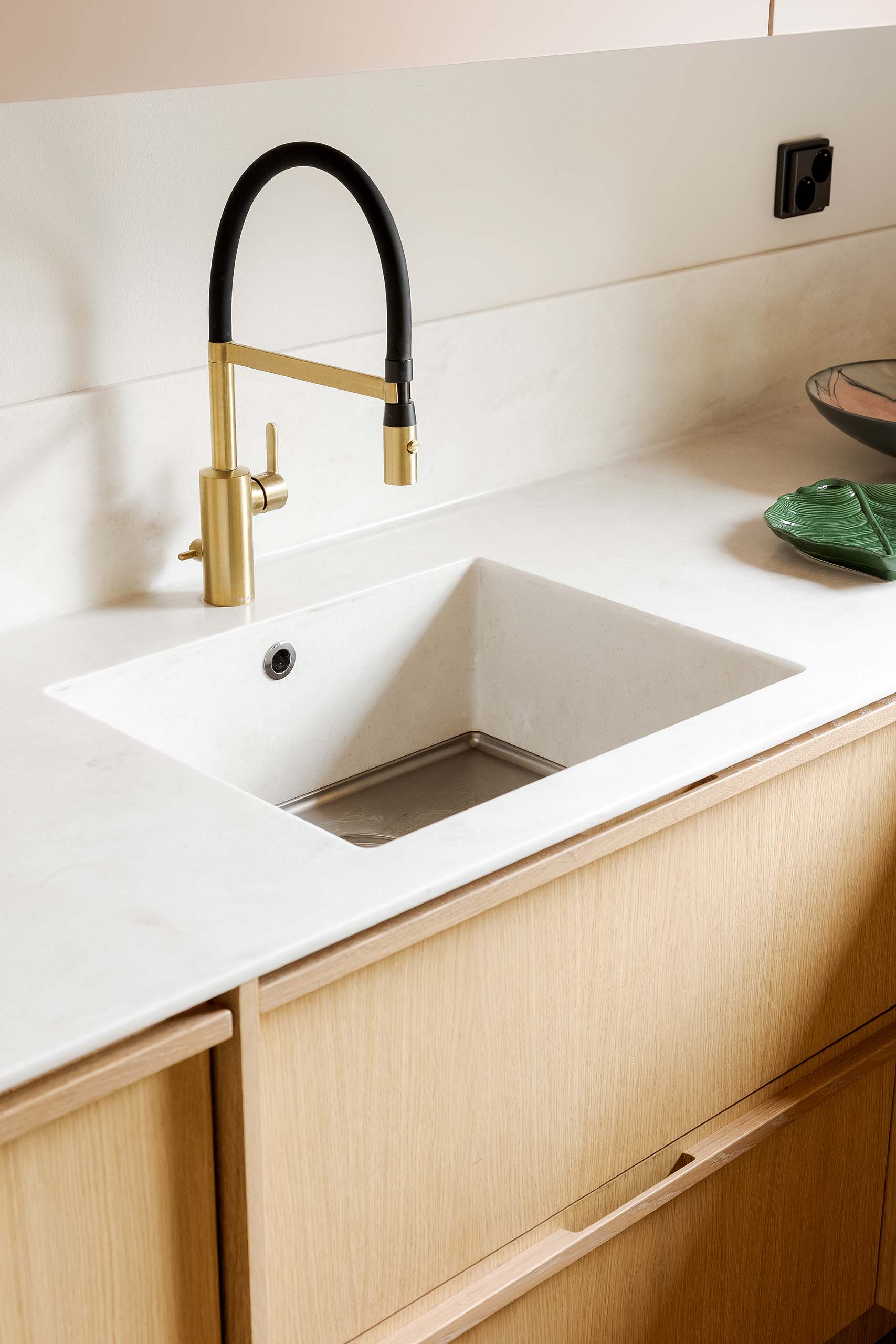 Modern kitchen sink with brass and black pull-down faucet set in white countertop and light wooden cabinets.