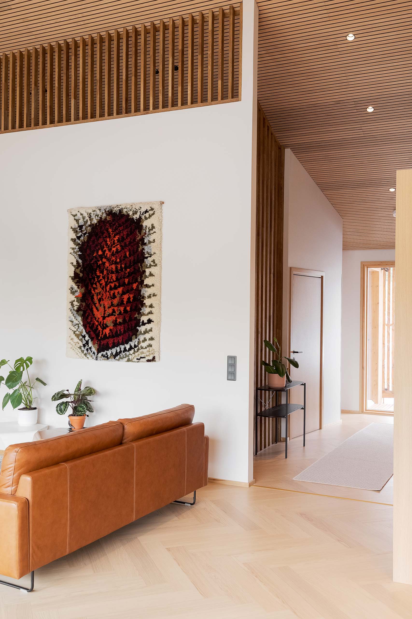 Modern living room with a tan leather sofa, two potted plants, a colorful woven wall hanging, and light wood flooring and ceiling with wooden slats.