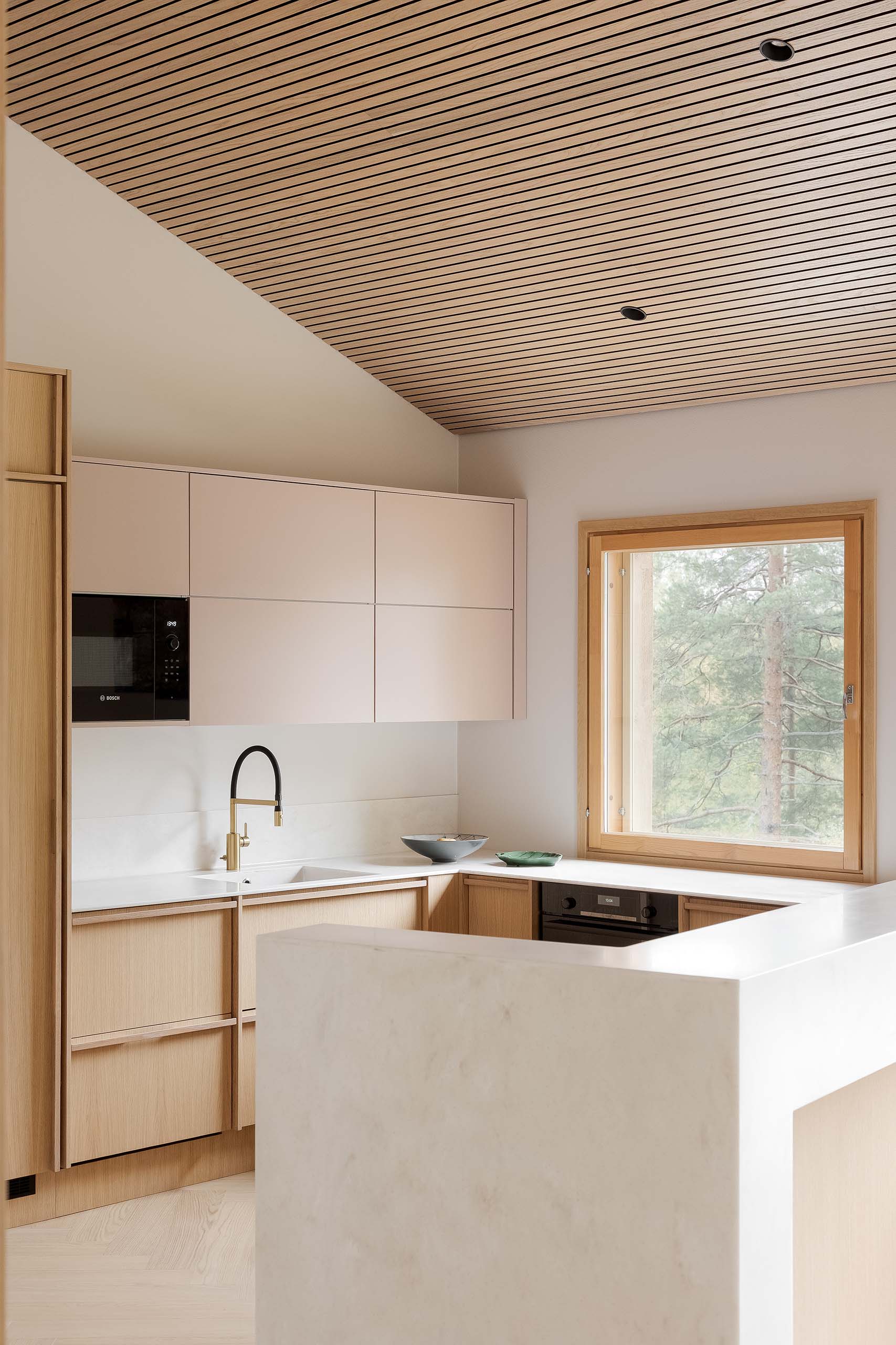 Modern kitchen with light wood cabinets, white countertops, black and gold faucet, black appliances, and a window overlooking trees.