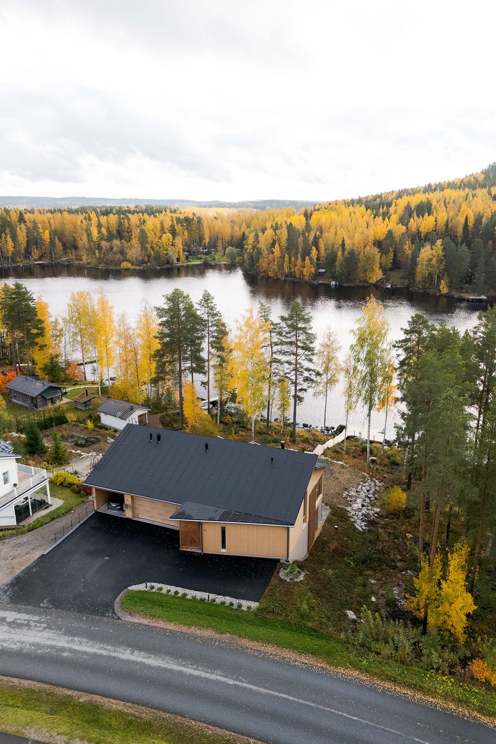 A modern house with a black roof next to a road, surrounded by autumn-colored trees and overlooking a lake with a dock.