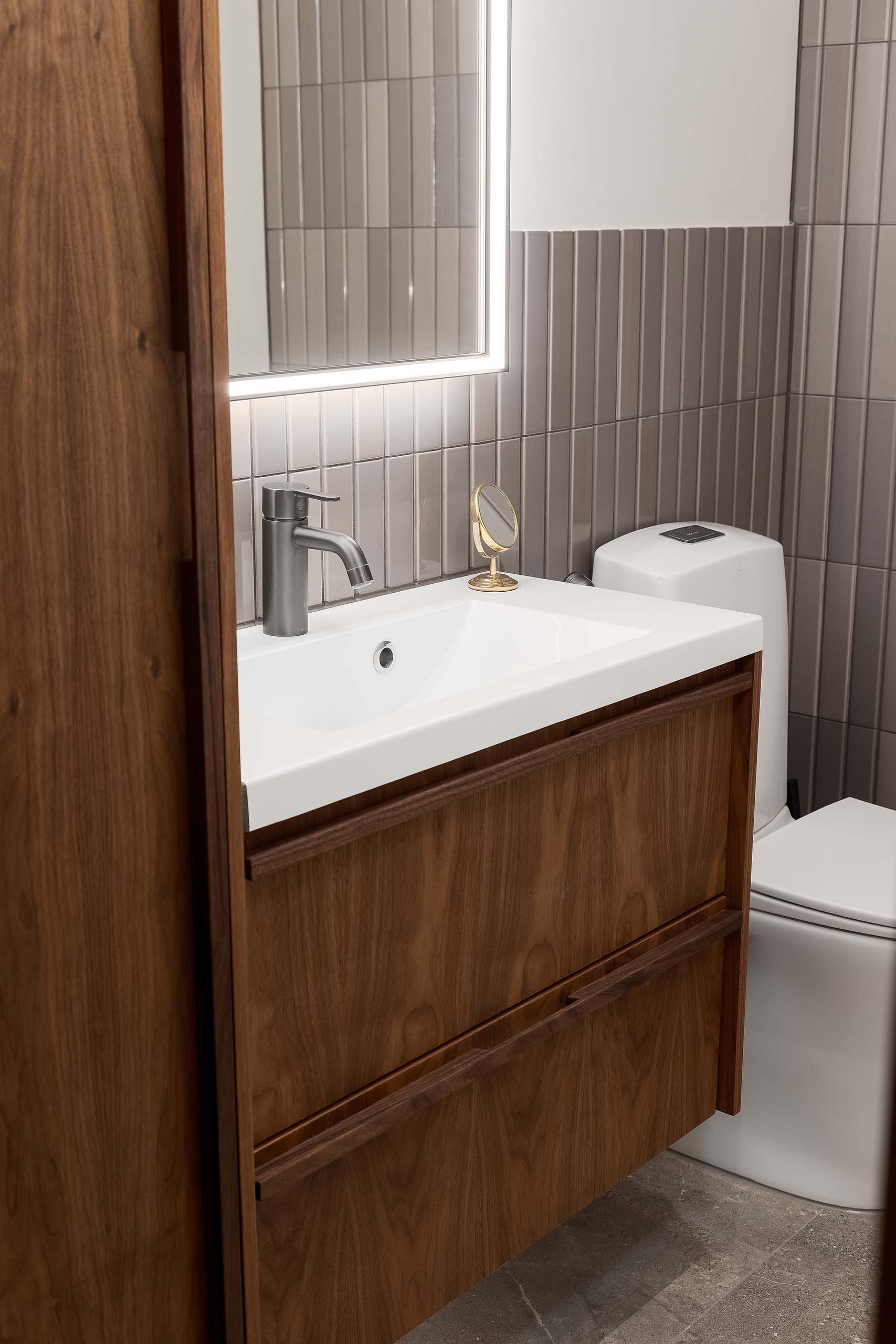 Modern bathroom sink with a wooden vanity, silver faucet, backlit mirror, and a white toilet with gray tiled walls.