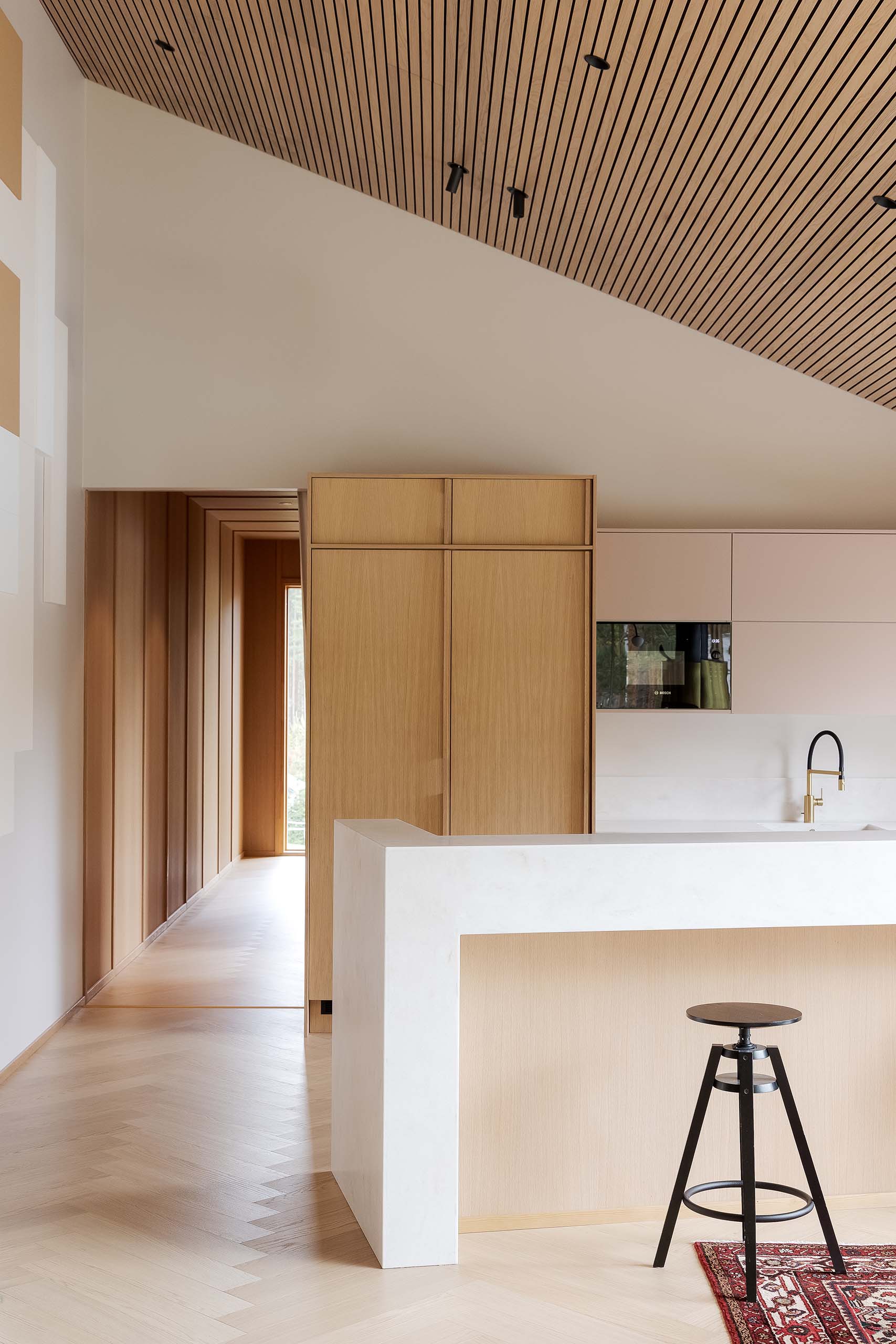 Minimalist kitchen with a white marble island, light wood cabinetry, black stool on a red patterned rug, and a slatted wood ceiling.