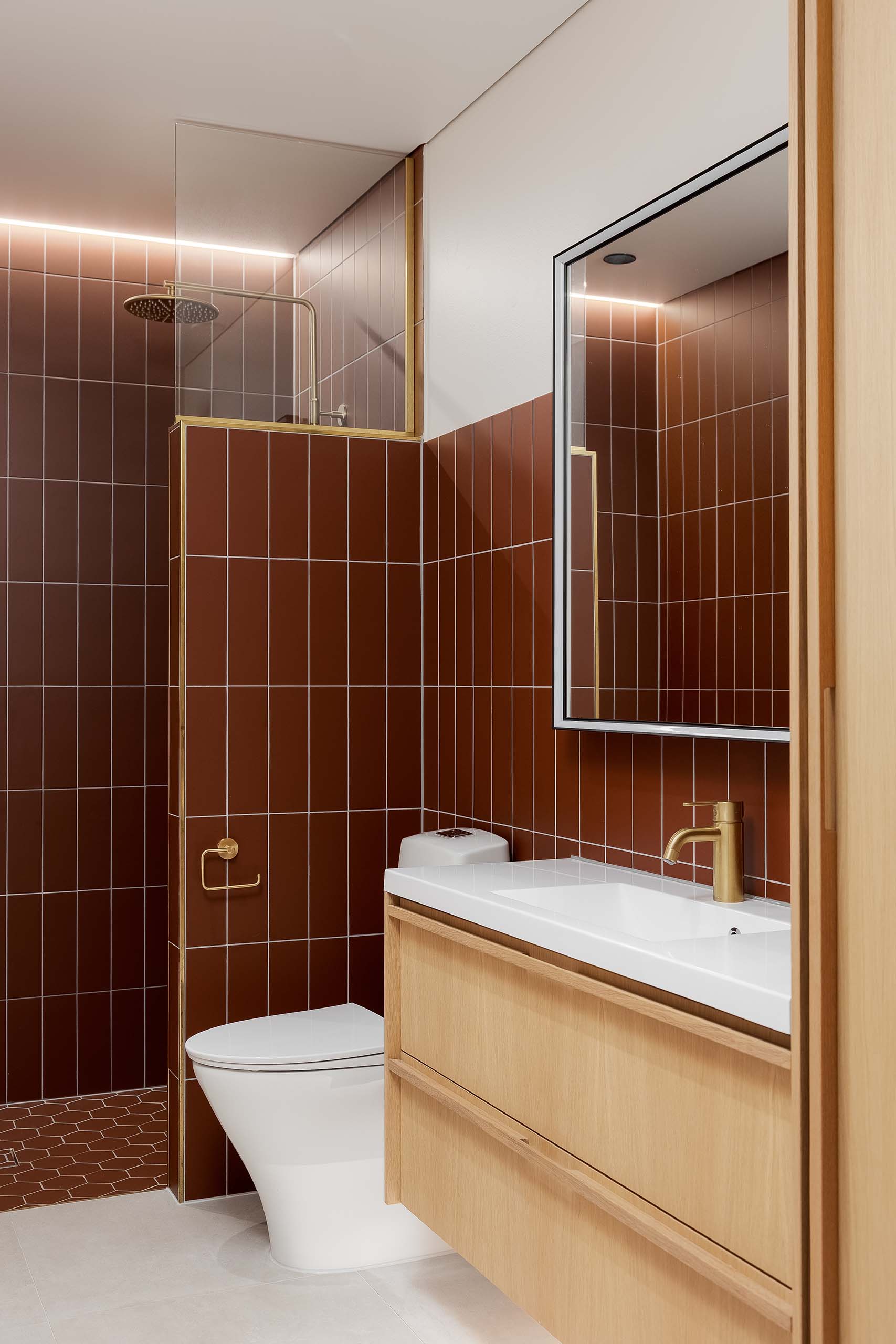 Modern bathroom with brown vertical tiles, a walk-in shower, white toilet, wooden vanity with white countertop, and gold faucet.