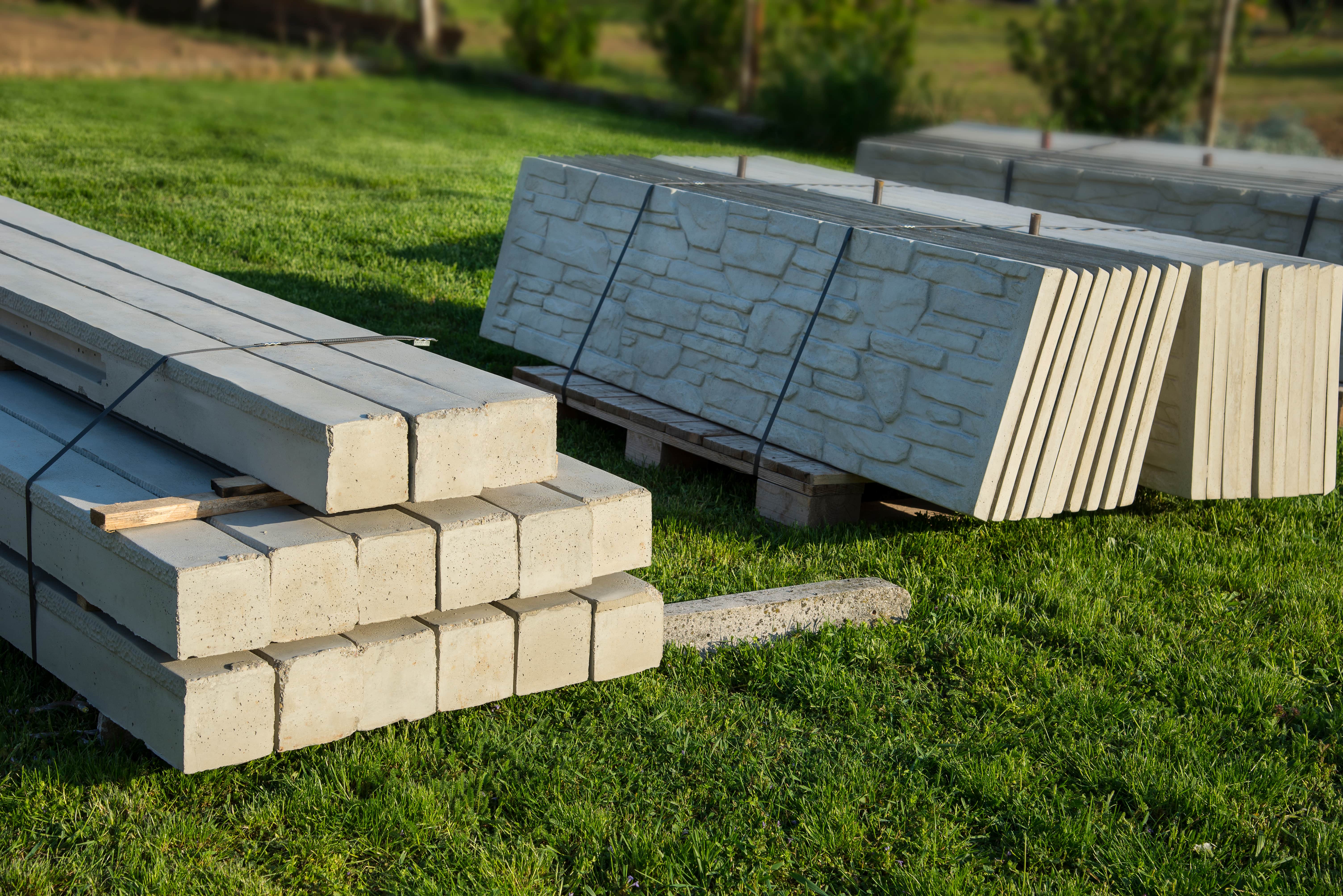 A ground-level view of stacks of precast concrete fence sections resting on pallets on a vibrant green lawn. The stack in the foreground contains smooth, rectangular posts, while the stacks behind it contain panels with a decorative, rock-like textured finish.