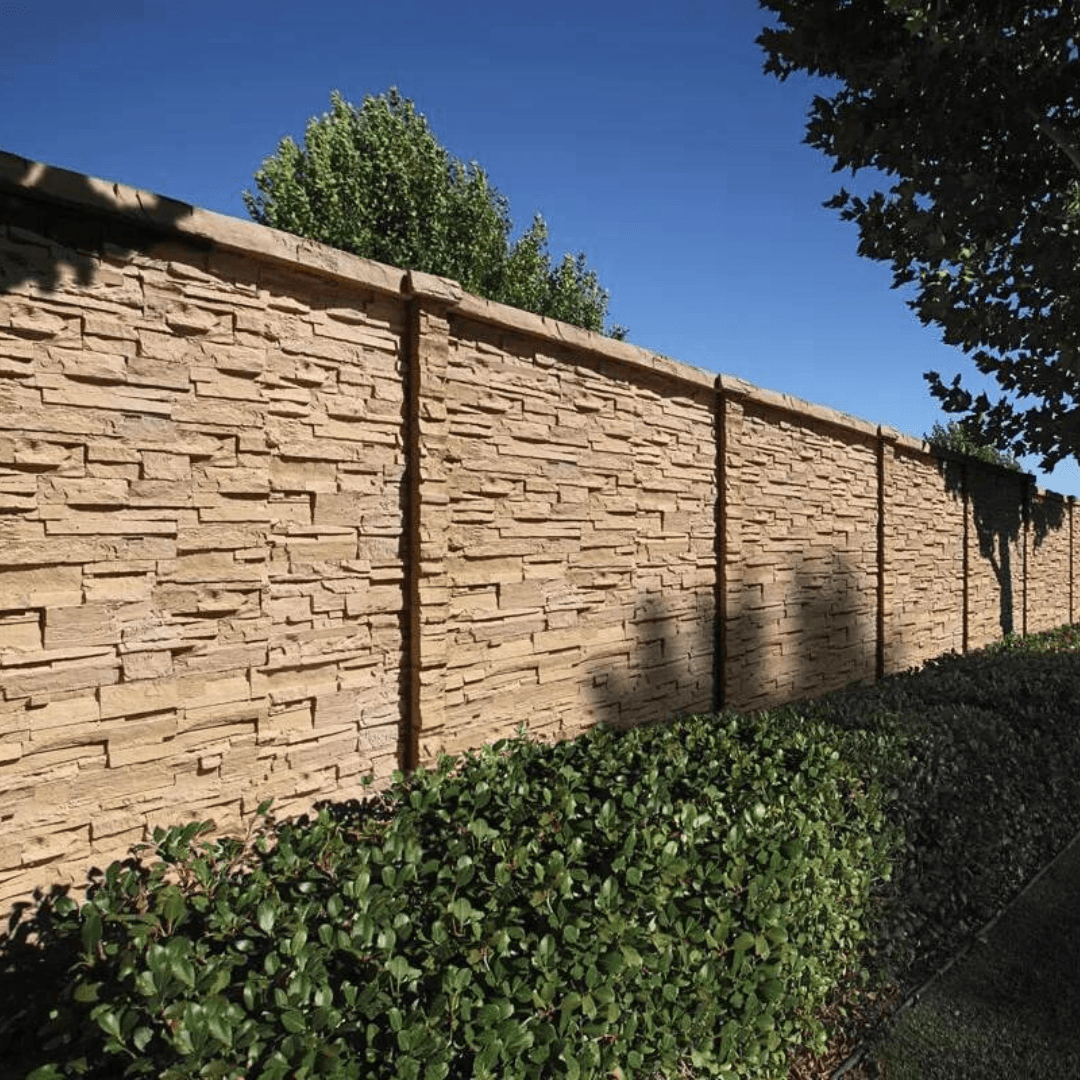 A long section of tall tan precast concrete wall with a stacked stone texture. The base is lined with dark green shrubs, and trees cast long shadows on the fence under a clear blue sky.
