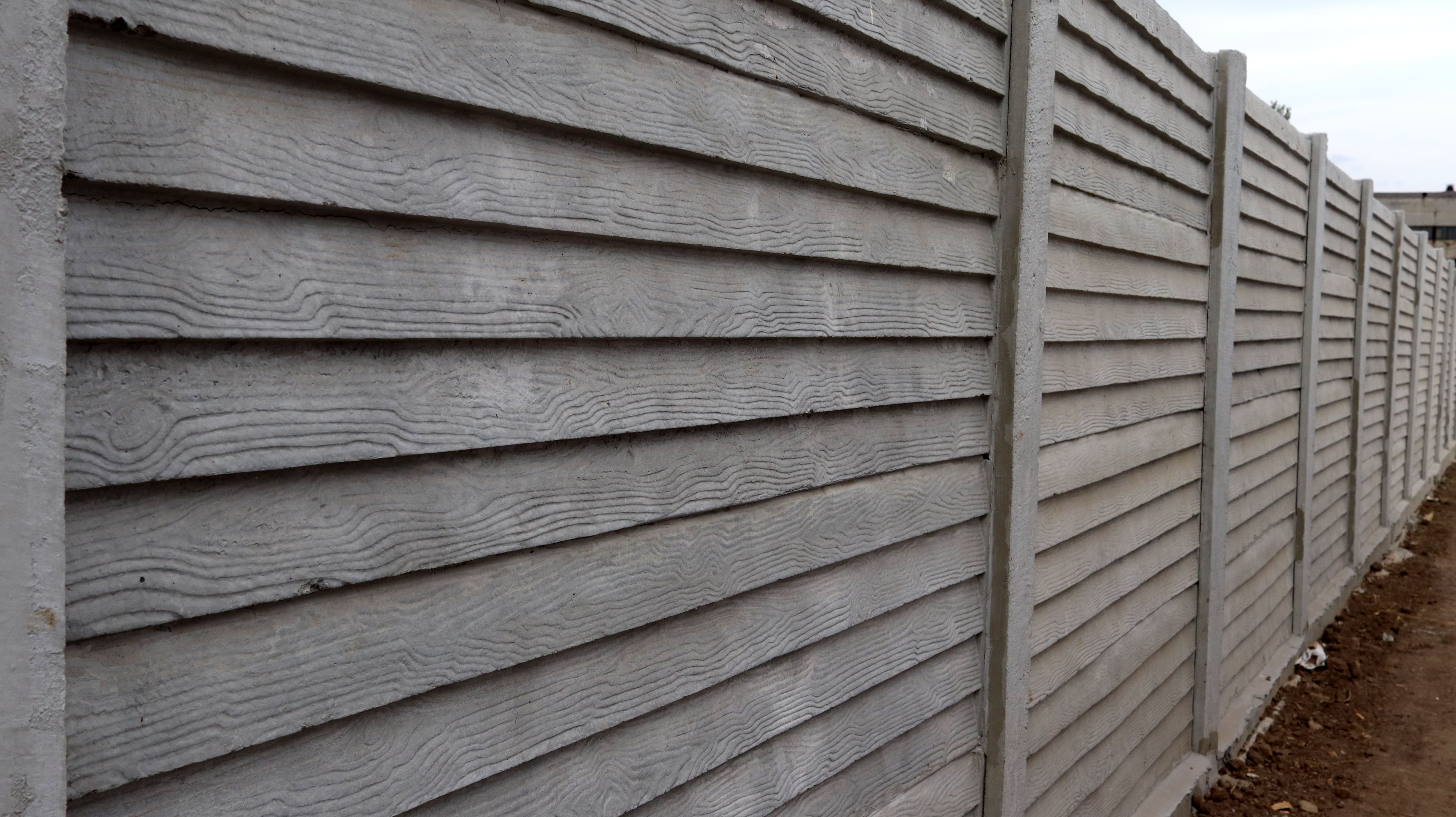 A long row of gray precast concrete fence panels textured to resemble horizontal wood siding, supported by concrete posts along a dirt path.