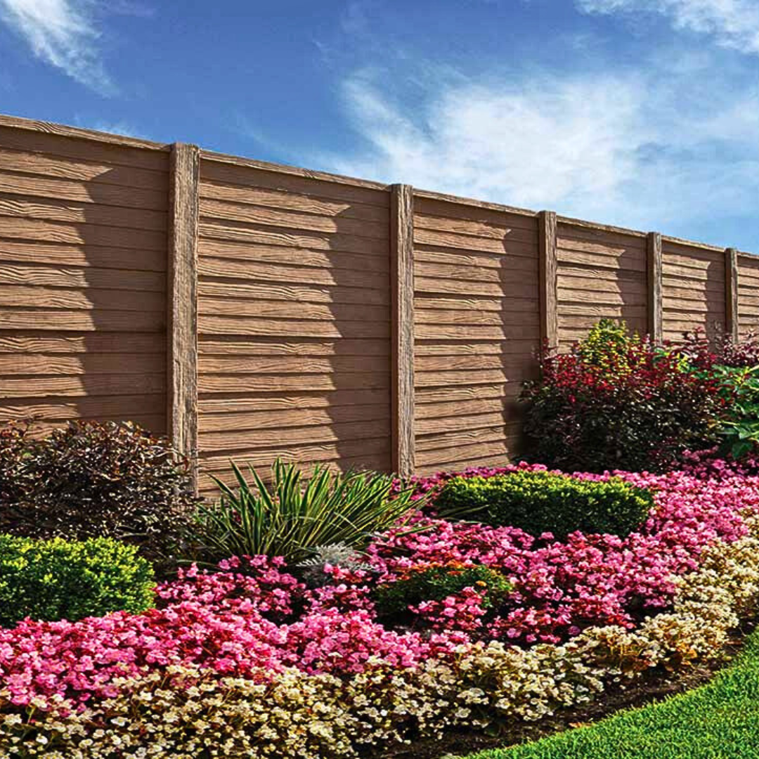 A tall, brown concrete fence textured to look like horizontal wood planks, bordering a lush garden bed of pink, red, and white flowers.